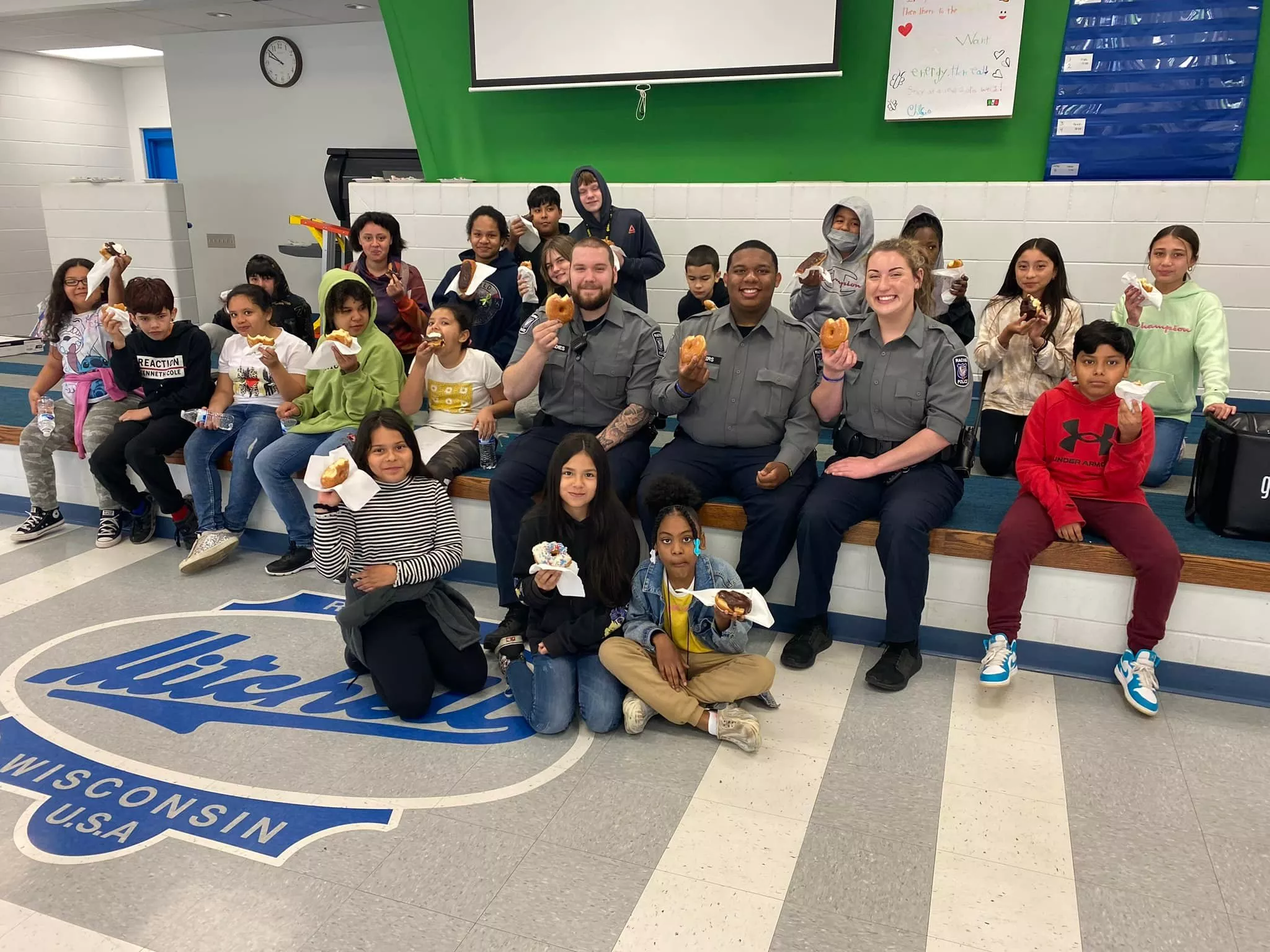 Group of students and officers holding food items, sitting on a school stage with the school logo.