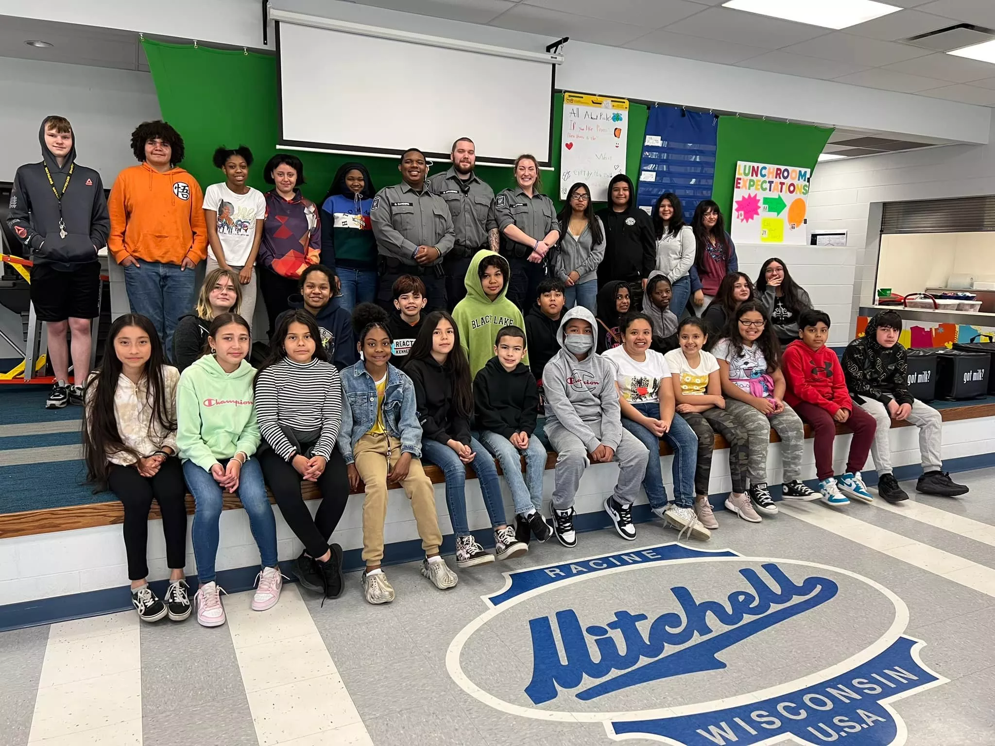 Group of students and officers pose in front of the Mitchell Elementary School logo.