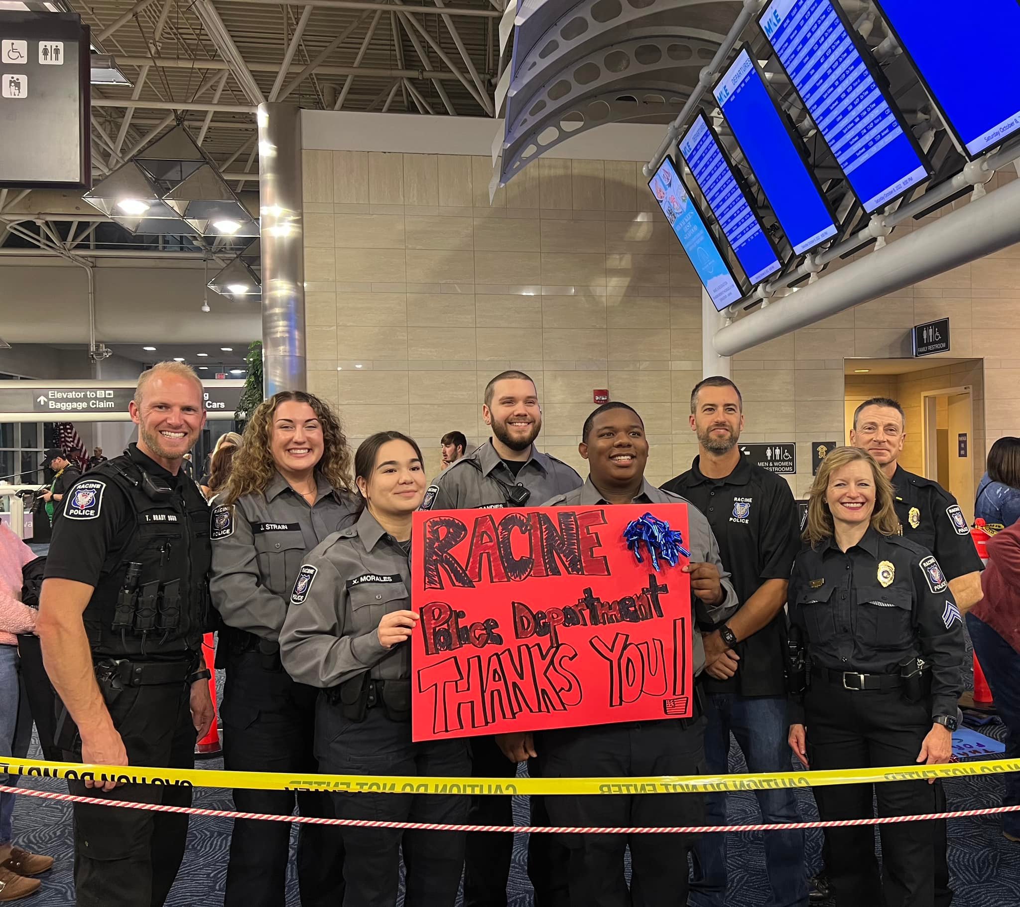 Group of police officers holding a thank you sign at an airport, with flight information boards in the background.