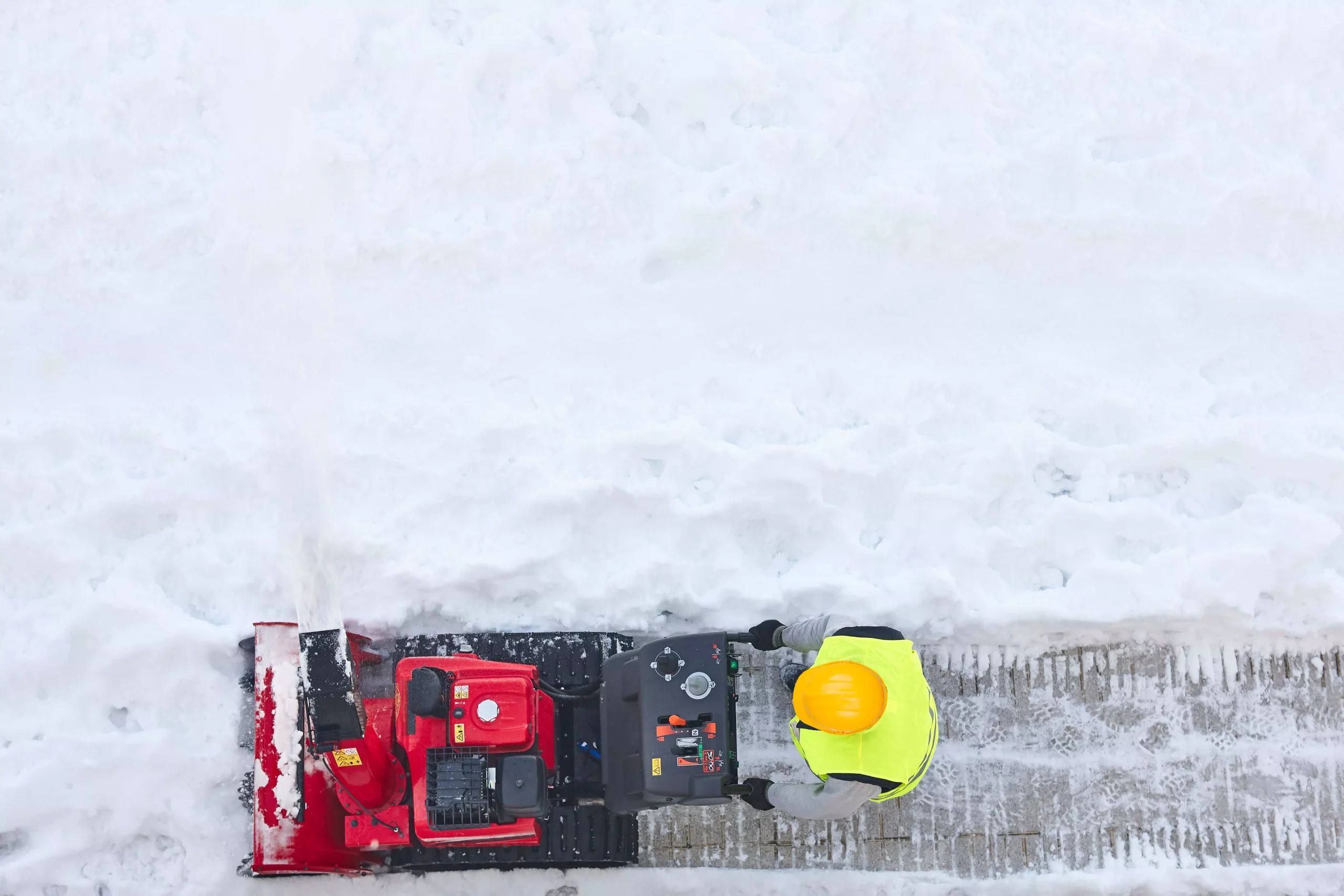 Overhead view of worker using red snowblower to clear sidewalk after snowfall. Worker wears yellow safety vest and hard hat.