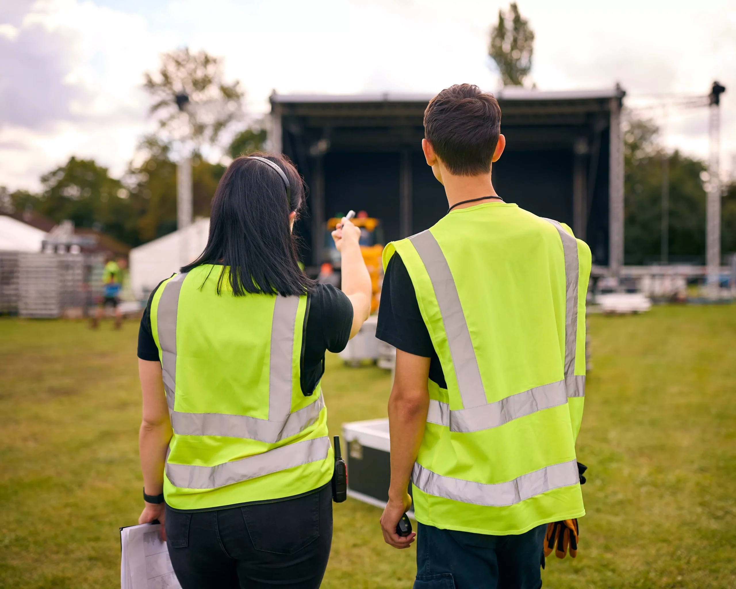 Two event staff in safety vests oversee stage setup on a grassy field. One points towards the stage.
