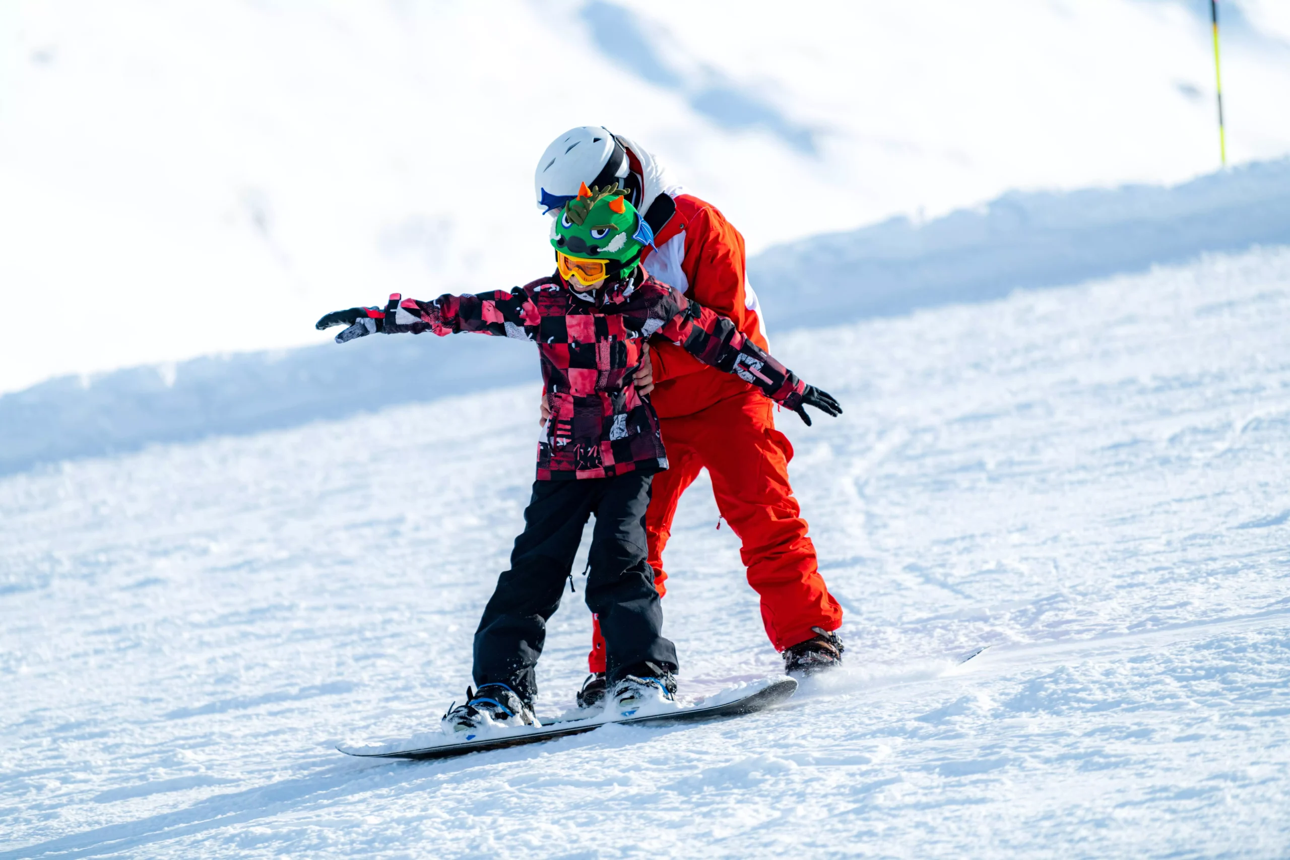 Child learning to snowboard with instructor on snowy slope, arms outstretched for balance.