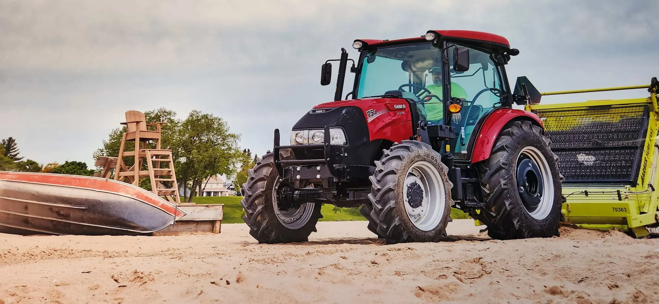 Red tractor with beach cleaner attachment on sand near lifeguard tower and overturned boat.