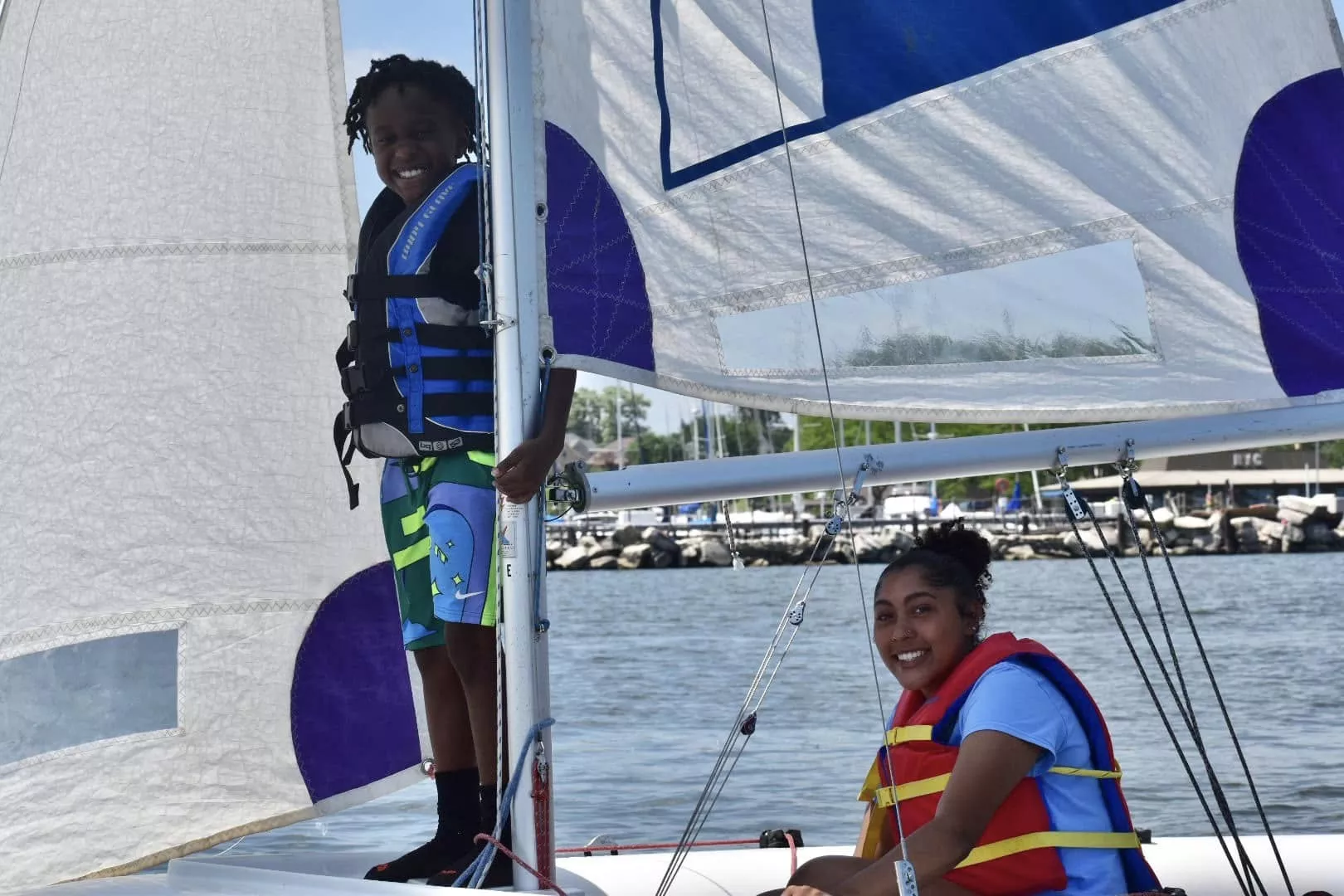 Two smiling young people sailing a small sailboat on the water, both wearing life vests.