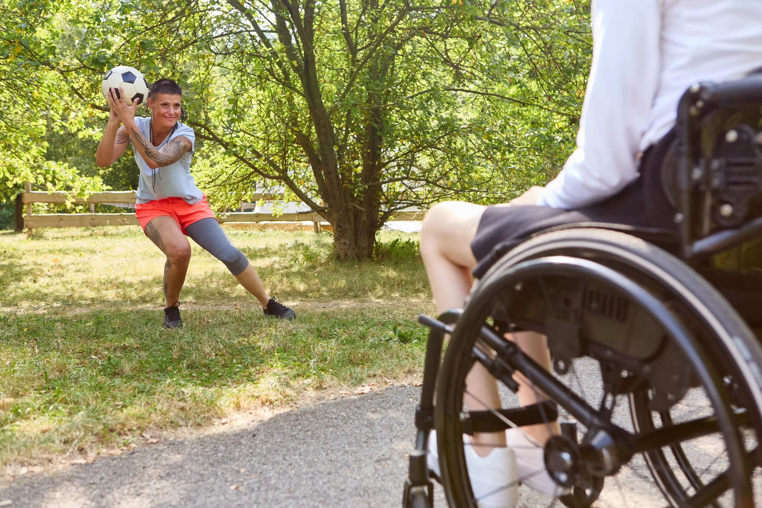 Woman throws soccer ball to person in wheelchair on a grassy path in a park setting.