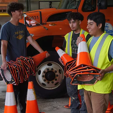 Three young men in safety vests holding stacks of orange traffic cones in front of a large orange truck.