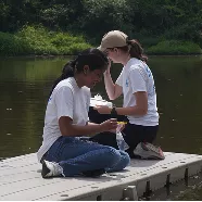 Two young women in t-shirts kneeling on a dock, examining water samples collected from the river.