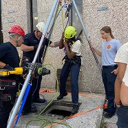 Rescue team assisting a person in a harness out of a confined space using a tripod and safety equipment.