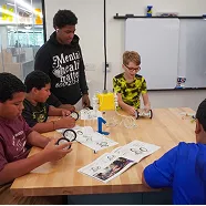 Group of children and a teen working on a robotics project around a table in a classroom setting.