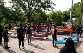 Group of people preparing to kayak, wearing life vests, with kayaks on the ground.
