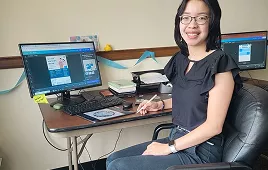 Young woman with glasses smiles, working on graphic design at her desk with two monitors and a drawing tablet.