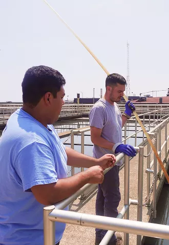 Two workers monitoring water quality at a wastewater treatment plant using a long sampling pole.
