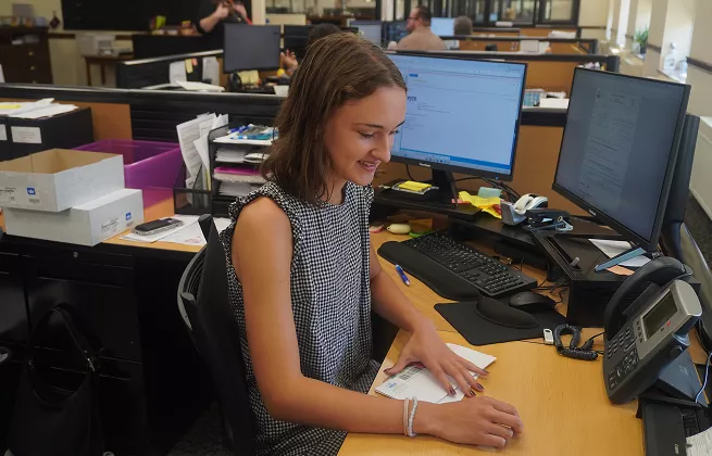Woman working at her desk in an office, smiling as she handles paperwork. Computer and phone are visible.