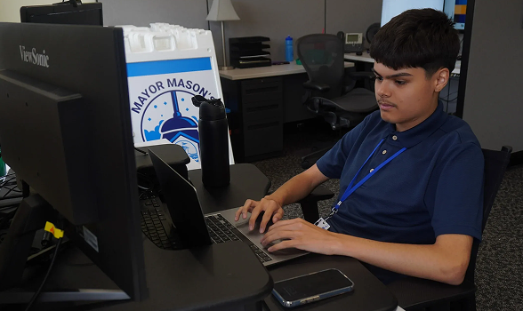 Teenage boy working on a laptop at a desk with a monitor and Mayor Mason's sign in the background.