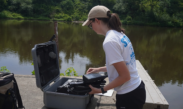 Scientist in hat and t-shirt examines equipment in open case on dock near water, trees in background.