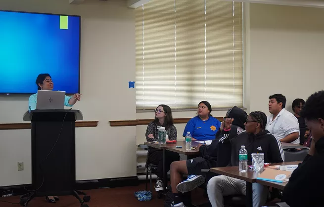 Presenter gestures towards screen while addressing seated audience in a classroom setting.