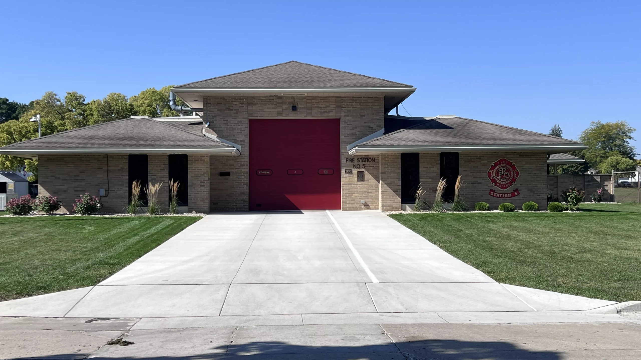 Exterior view of the brick fire station with a red garage door and manicured lawn on a sunny day.