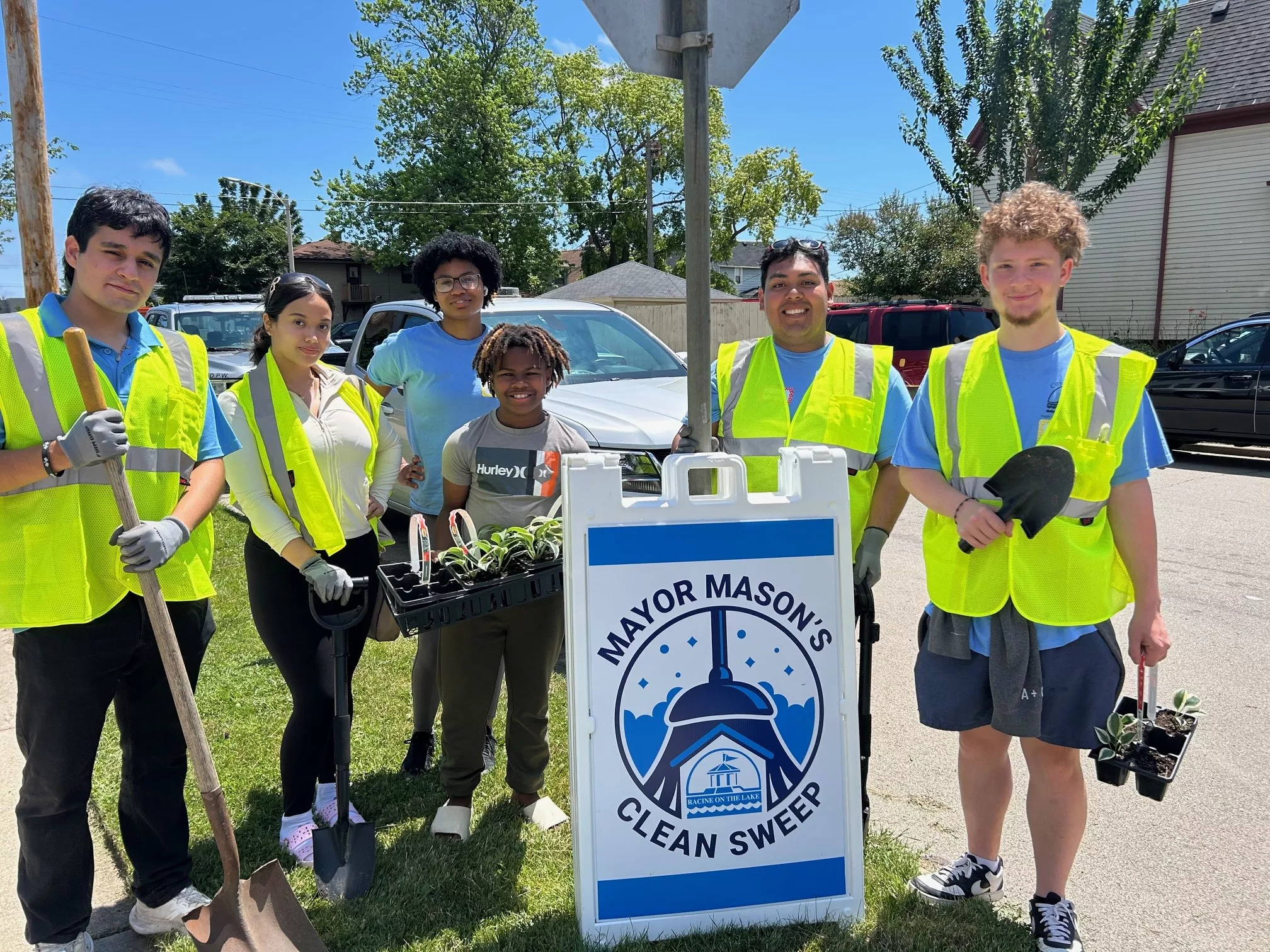 Group of volunteers wearing safety vests, holding gardening tools and plants next to a 'Mayor Mason's Clean Sweep' sign.