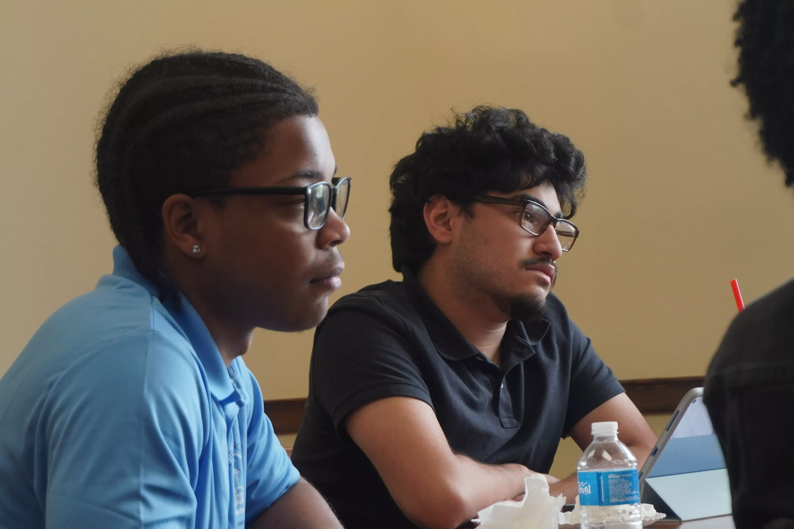 Two young men wearing glasses sit at a table, looking attentively at something off-screen. A water bottle is visible.