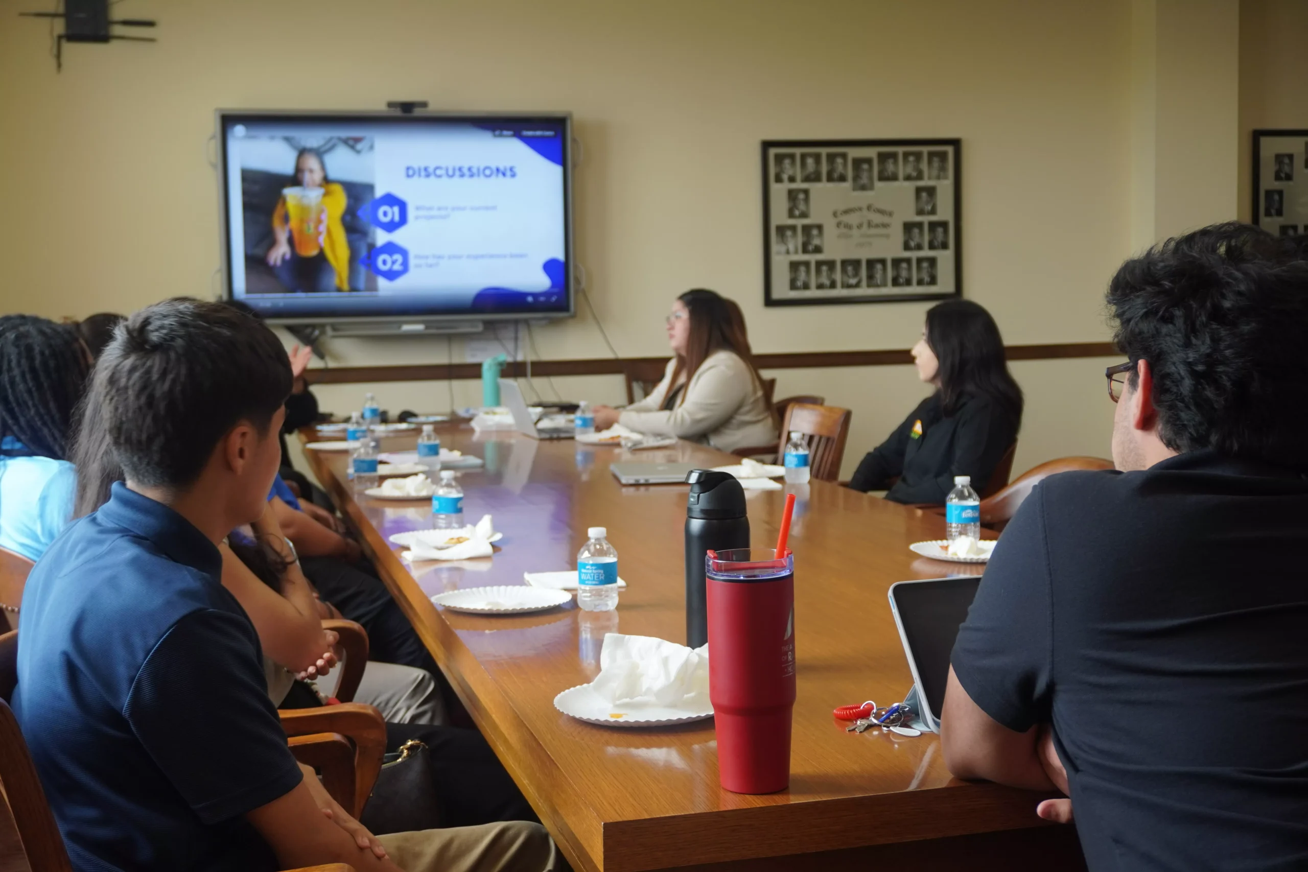 Group of people sit around a table watching a presentation on a large screen during a meeting.