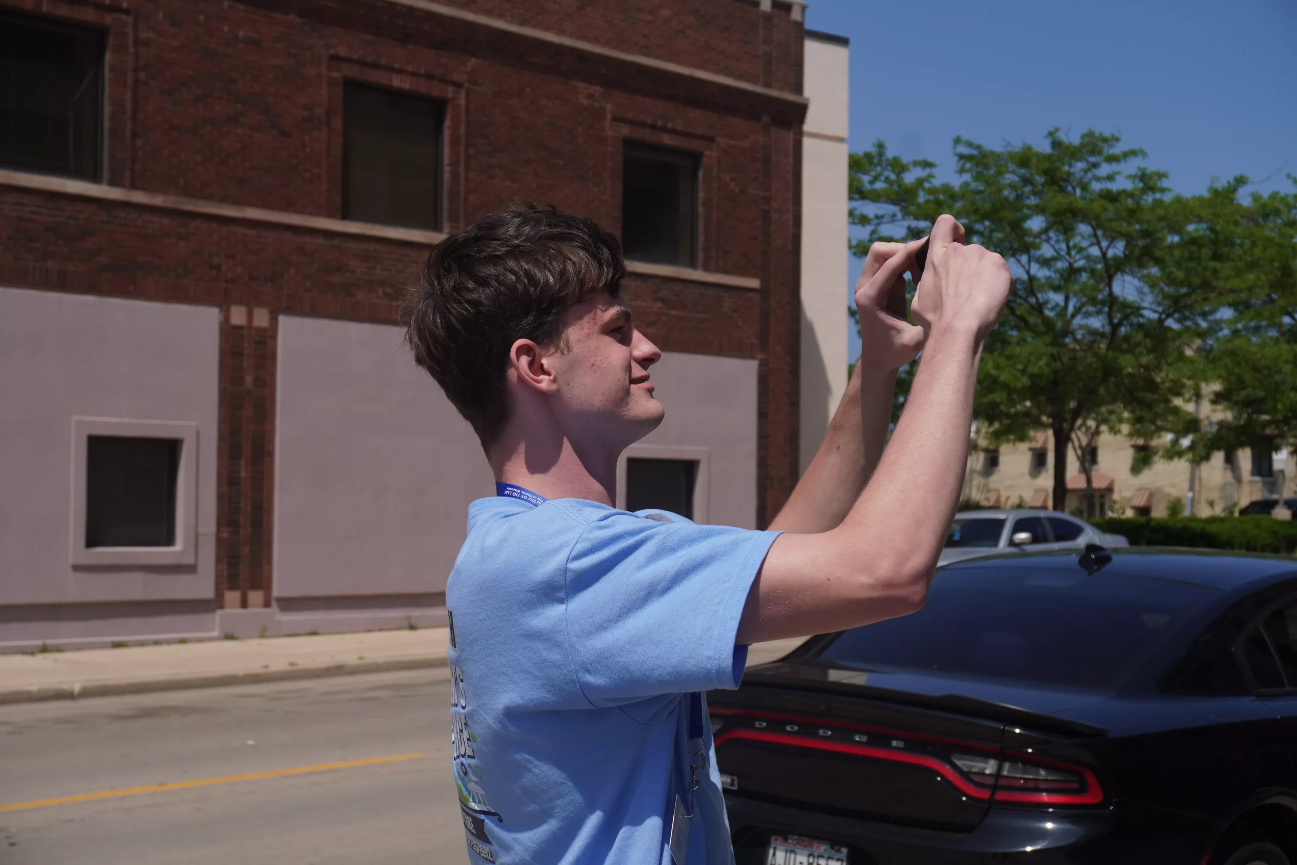 Young man taking a photo with his phone outdoors on a sunny day. He is wearing a blue shirt.