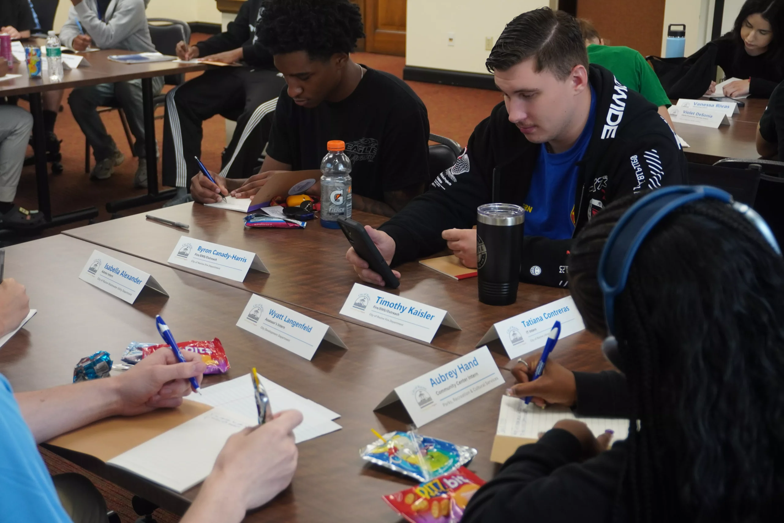 Students seated at a table, writing during a workshop or class. Name cards are visible on the table.