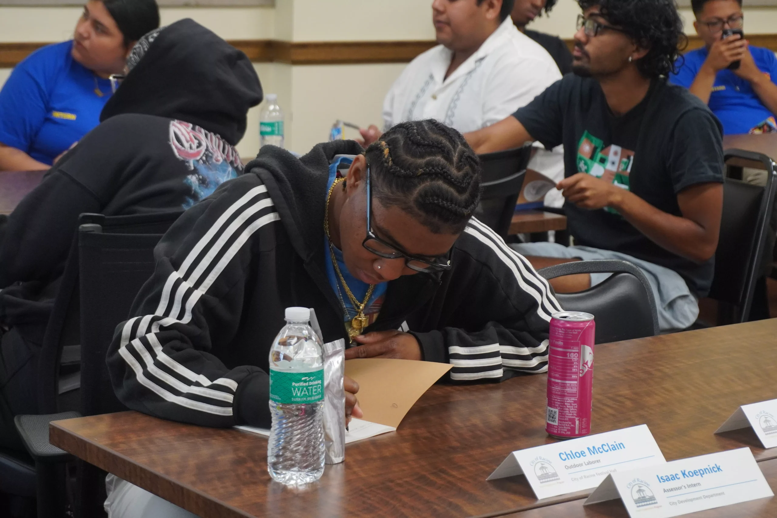 Student with braided hair focuses on a document at a table with water and a can of soda.