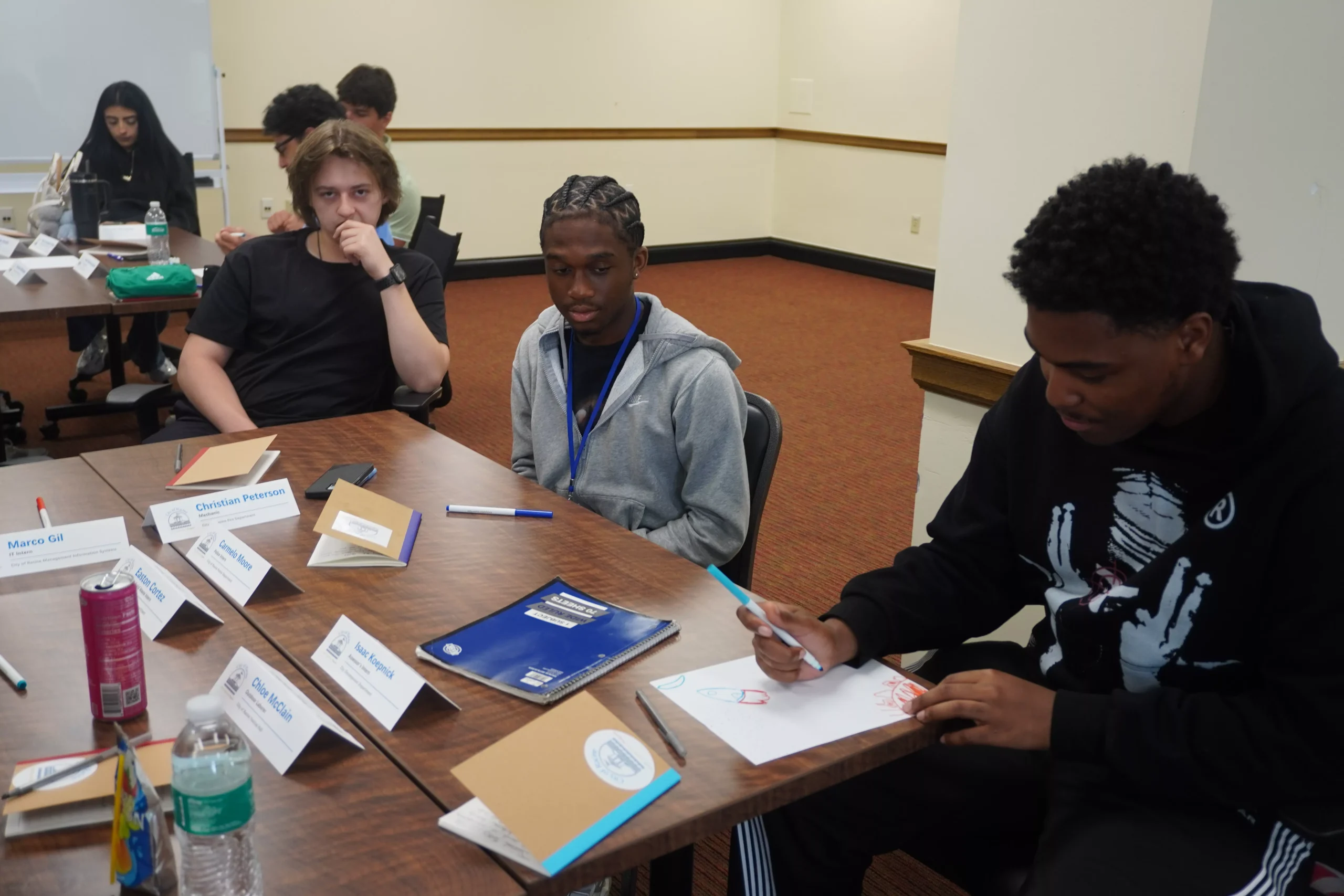 Students sitting at a table, one drawing on paper, others looking on in a classroom setting.