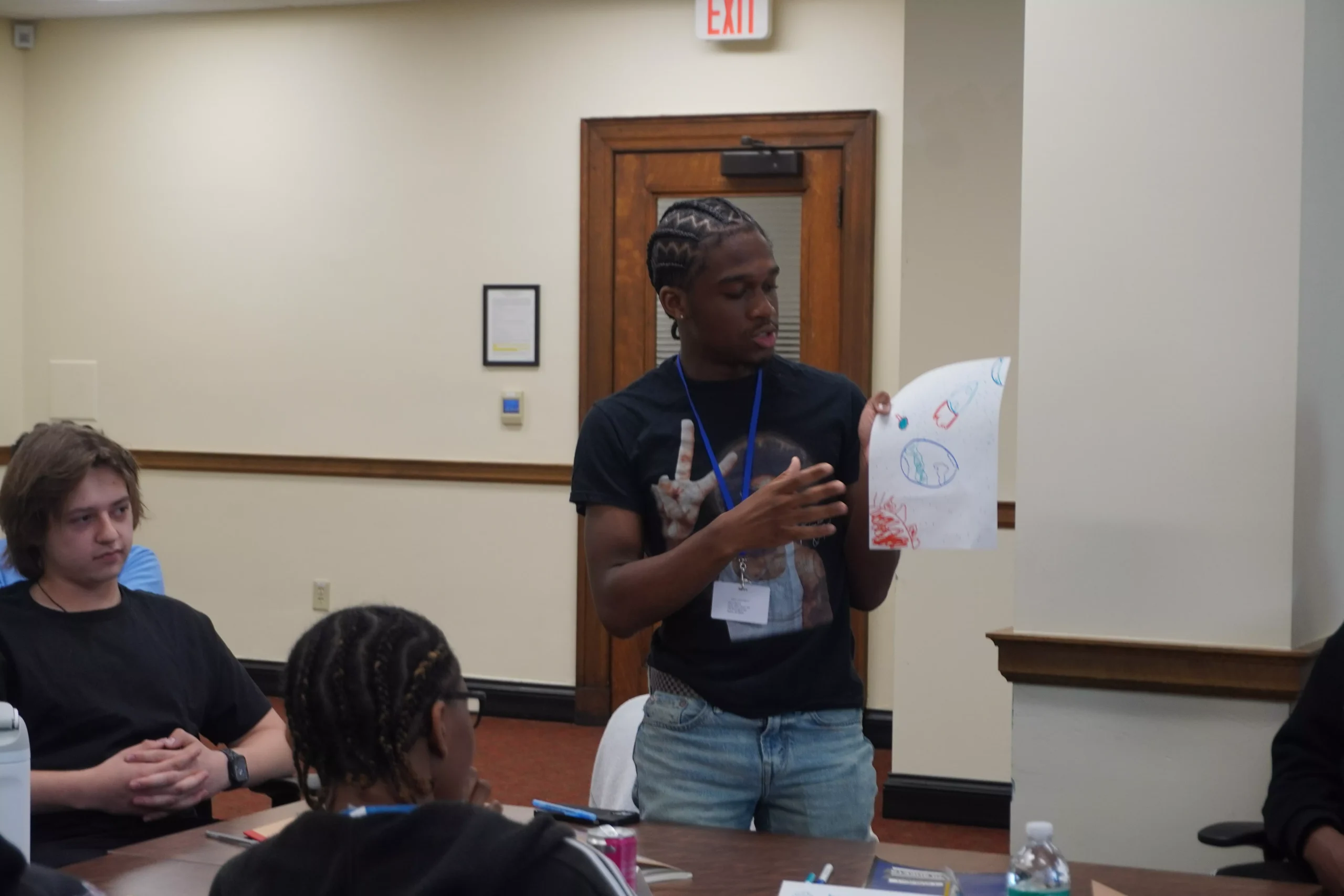 Person presenting a drawing to a group in a meeting room. The drawing features simple shapes and colors.