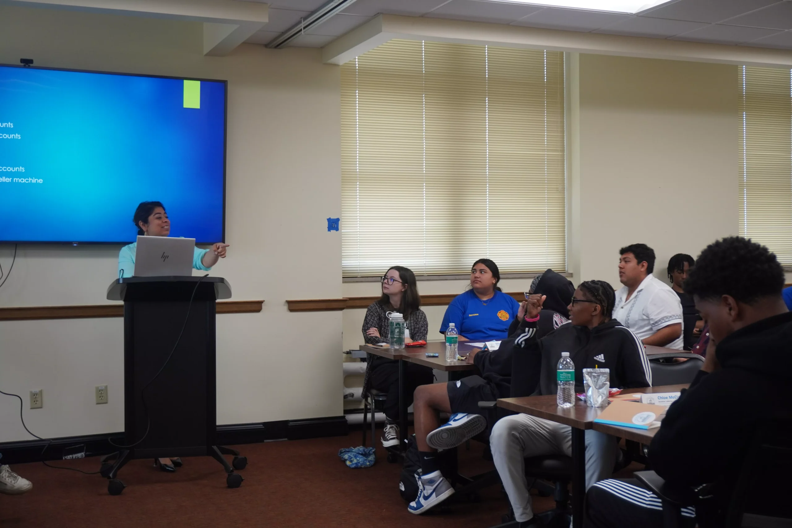 Presenter at podium addresses diverse audience in classroom with presentation screen.