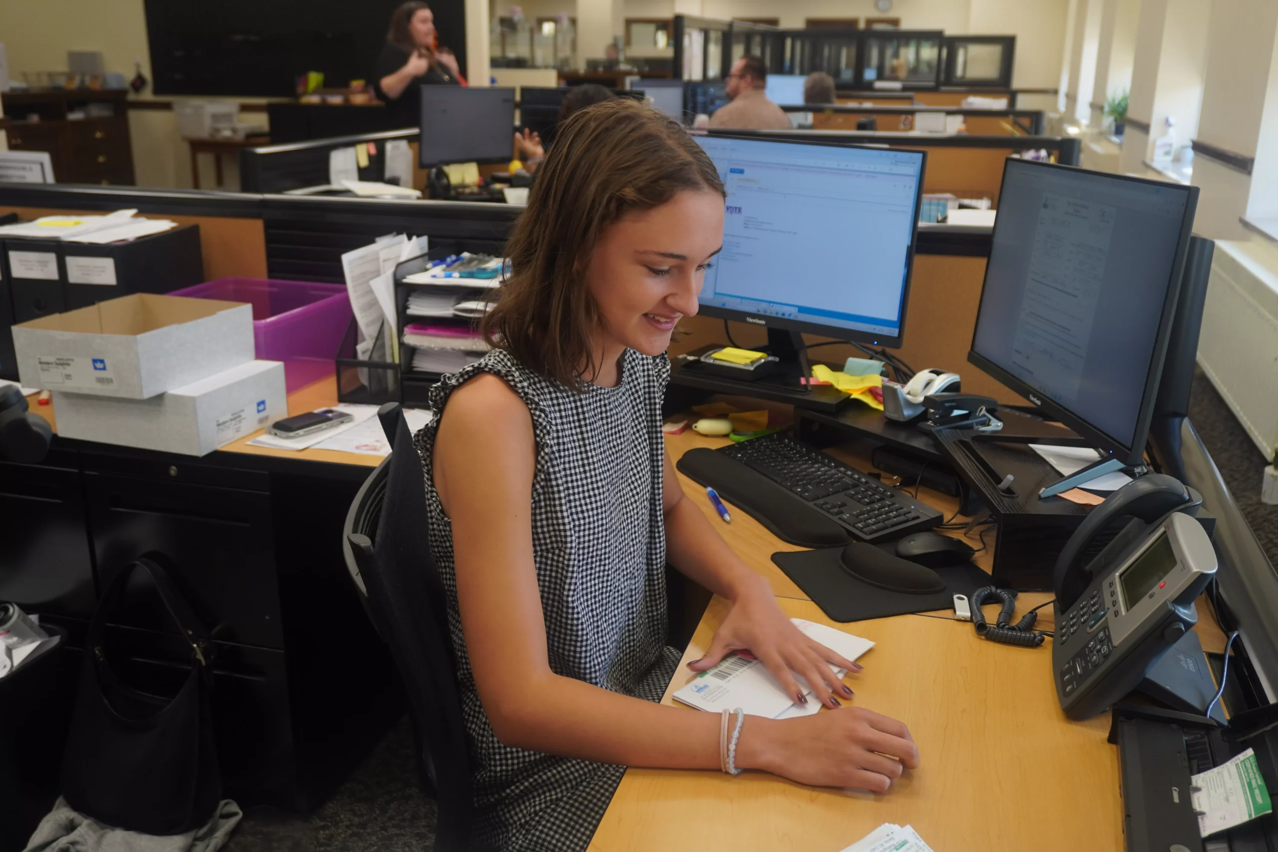 Woman working at a desk in an office, reviewing documents and using a computer. Other employees are visible in the