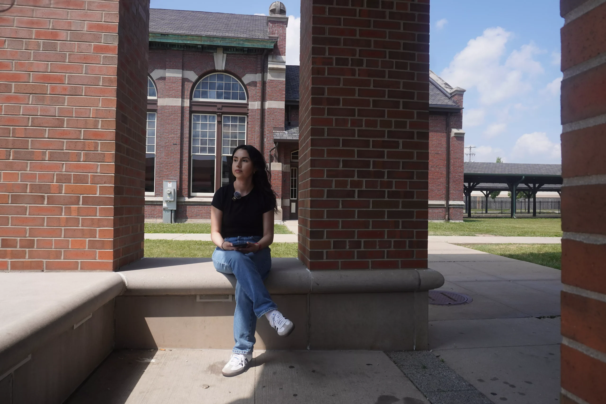 Woman sits on ledge outside brick building, holding phone, looking up and to the left.