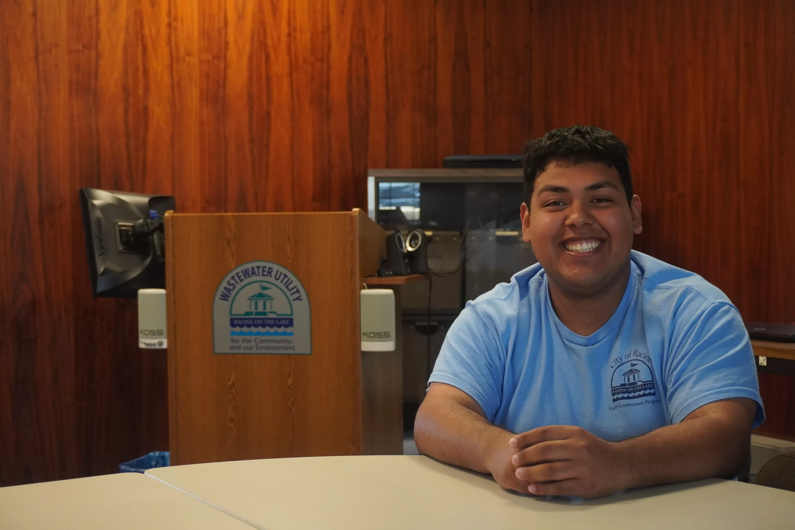 Smiling man in blue shirt sits at table next to Wastewater Utility podium. Wood paneled wall behind.