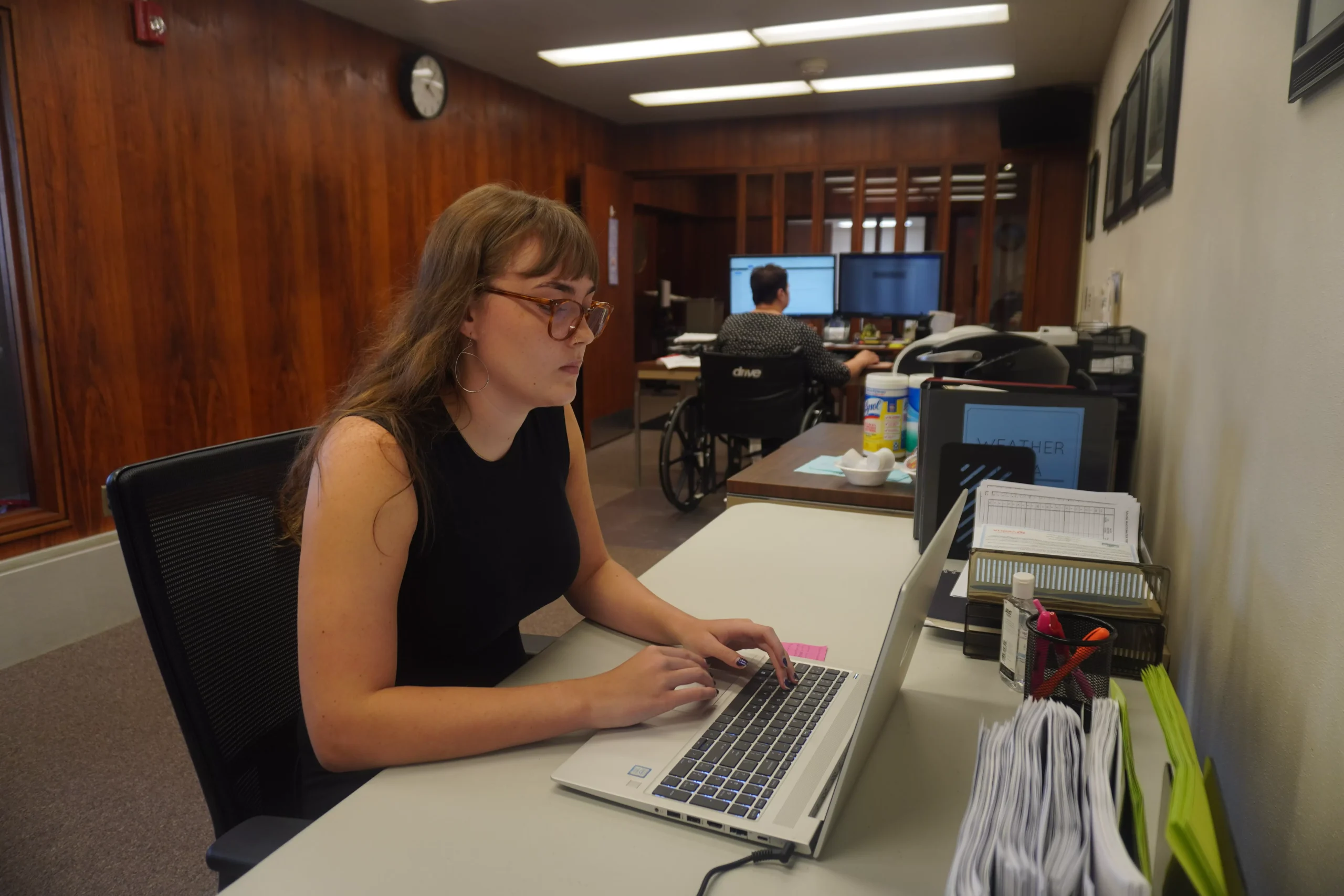 Woman with glasses types on laptop at desk in office; another person in wheelchair visible in background.