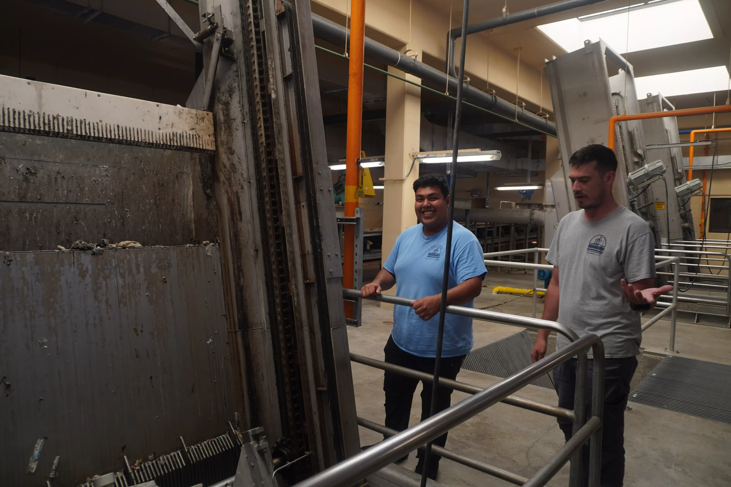 Two people stand near a large industrial machine inside a water treatment facility, likely during a tour or inspection.
