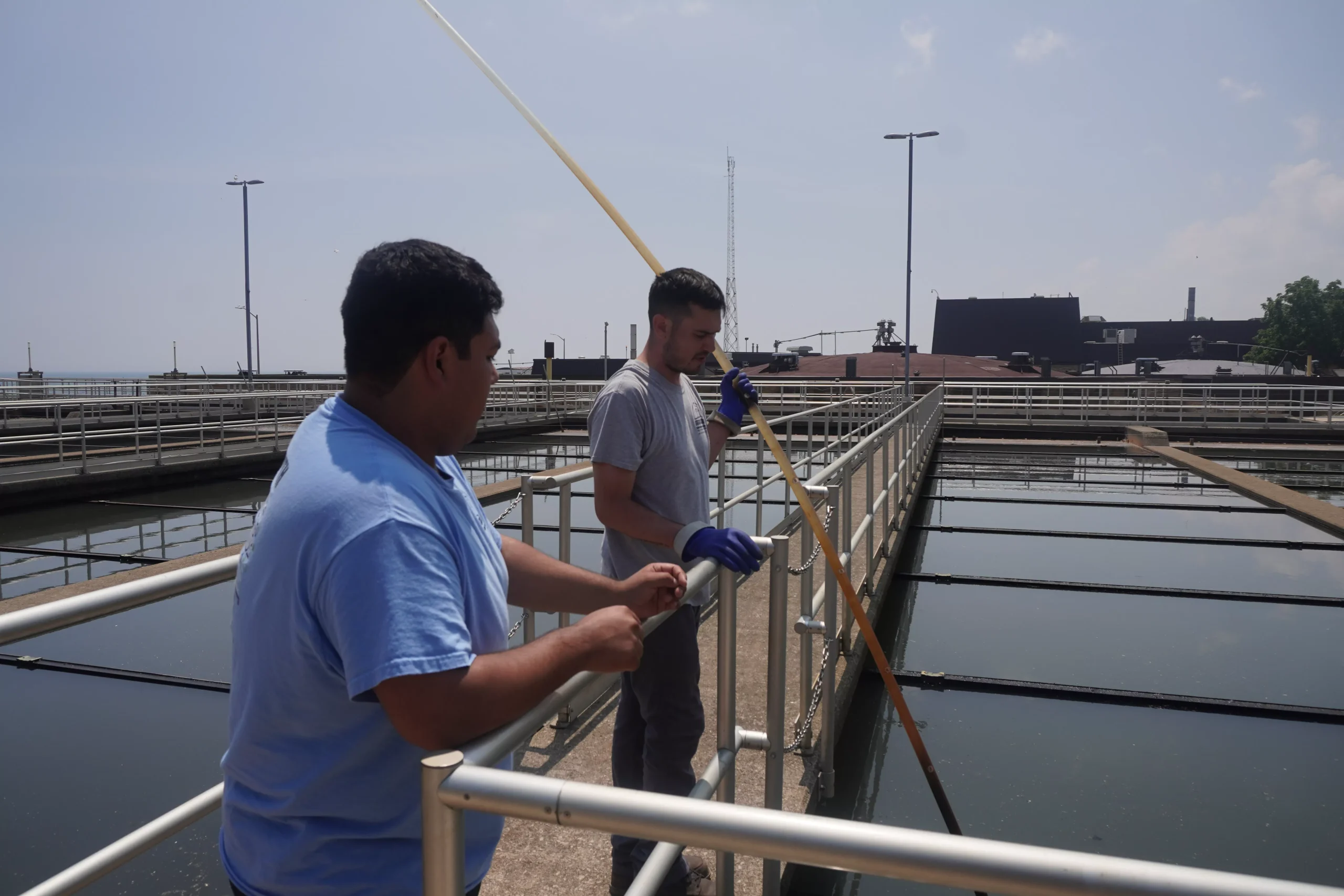 Two workers collect water samples at a water treatment plant, using a long pole to reach the water.