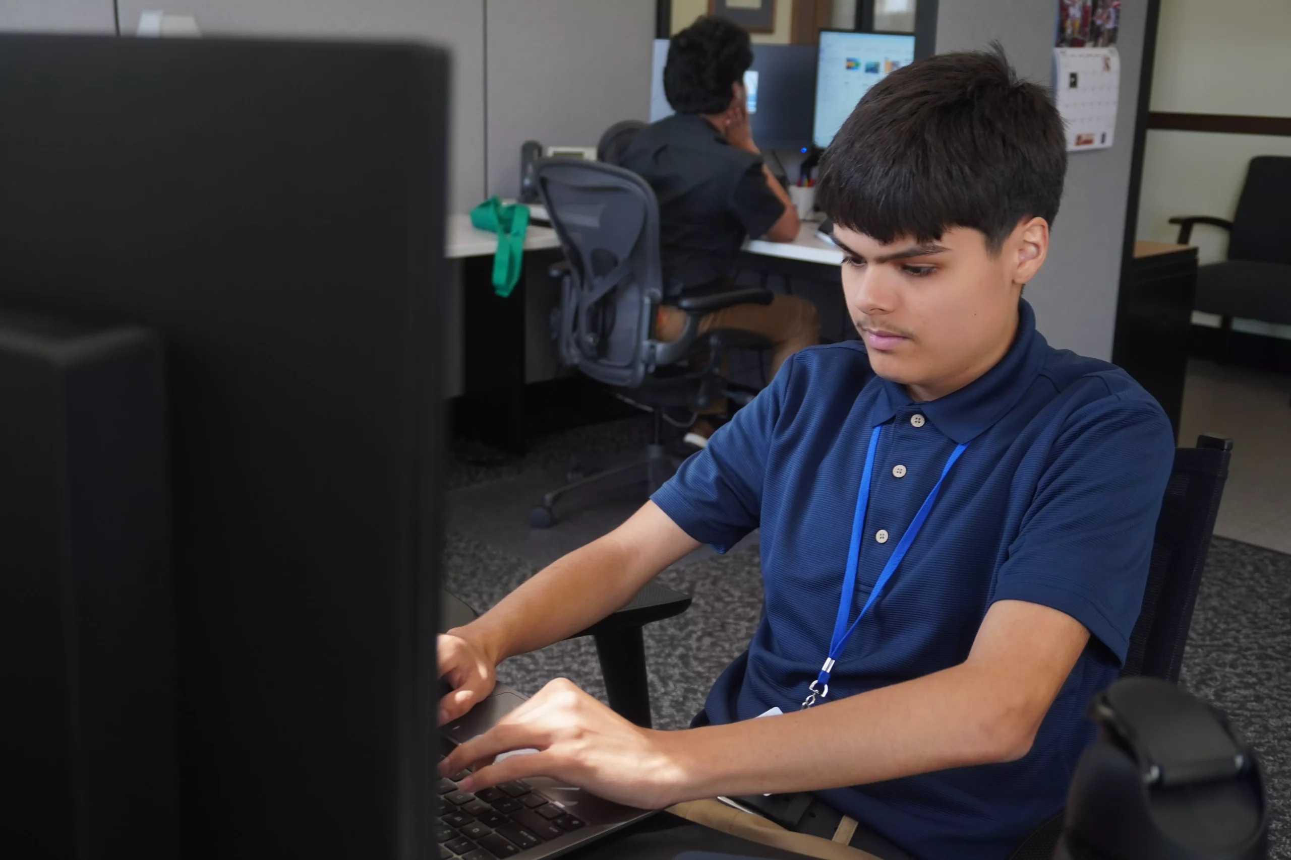 Young man working on a computer in an office setting, wearing a blue shirt and lanyard.