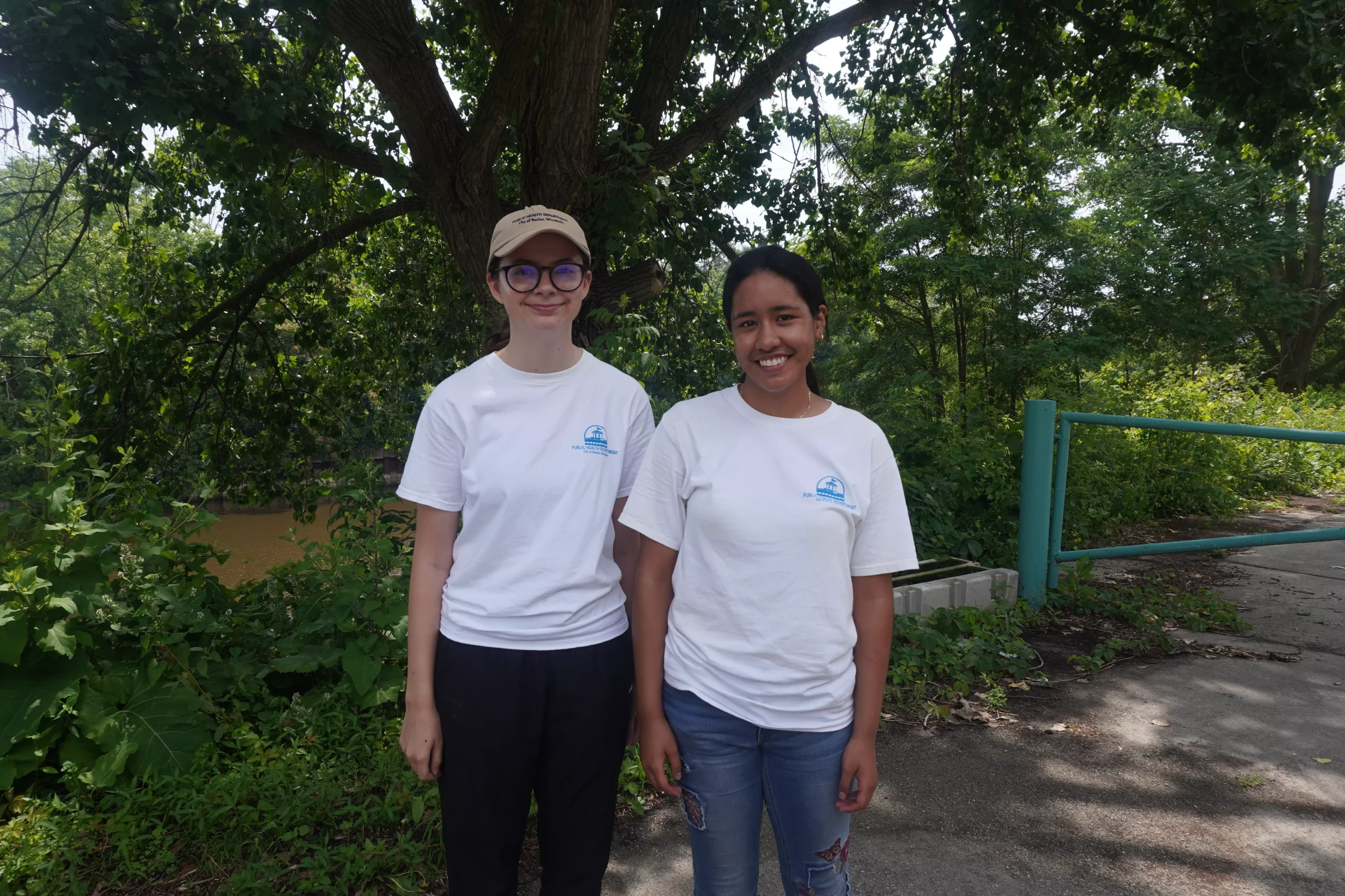 Two women wearing matching t-shirts stand outdoors near a river and trees, possibly volunteering for an environmental cause.