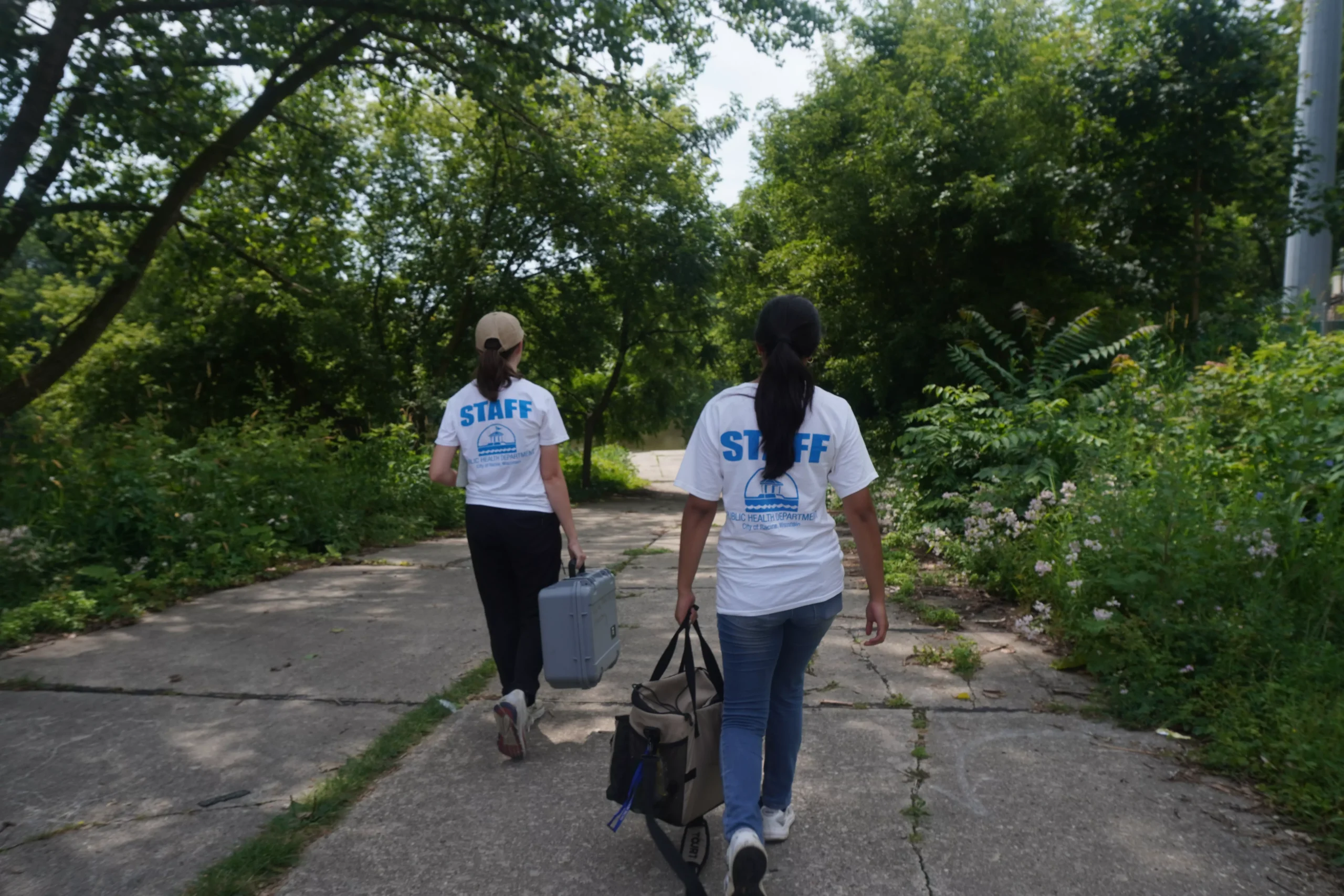 Two staff members walk on a path carrying equipment, with trees in the background.