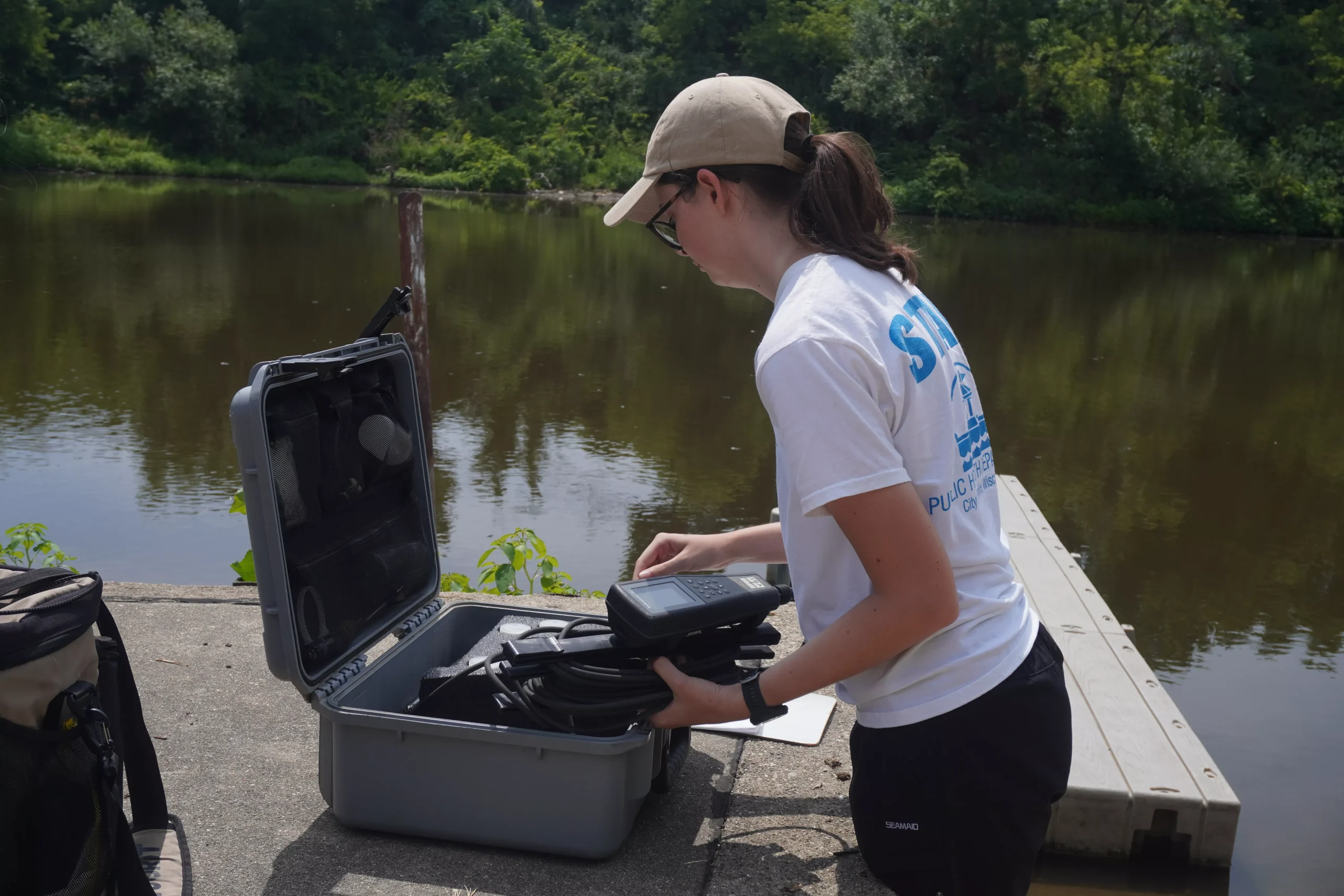 Scientist in field gear prepares equipment on a dock near a river, wearing a hat and uniform.