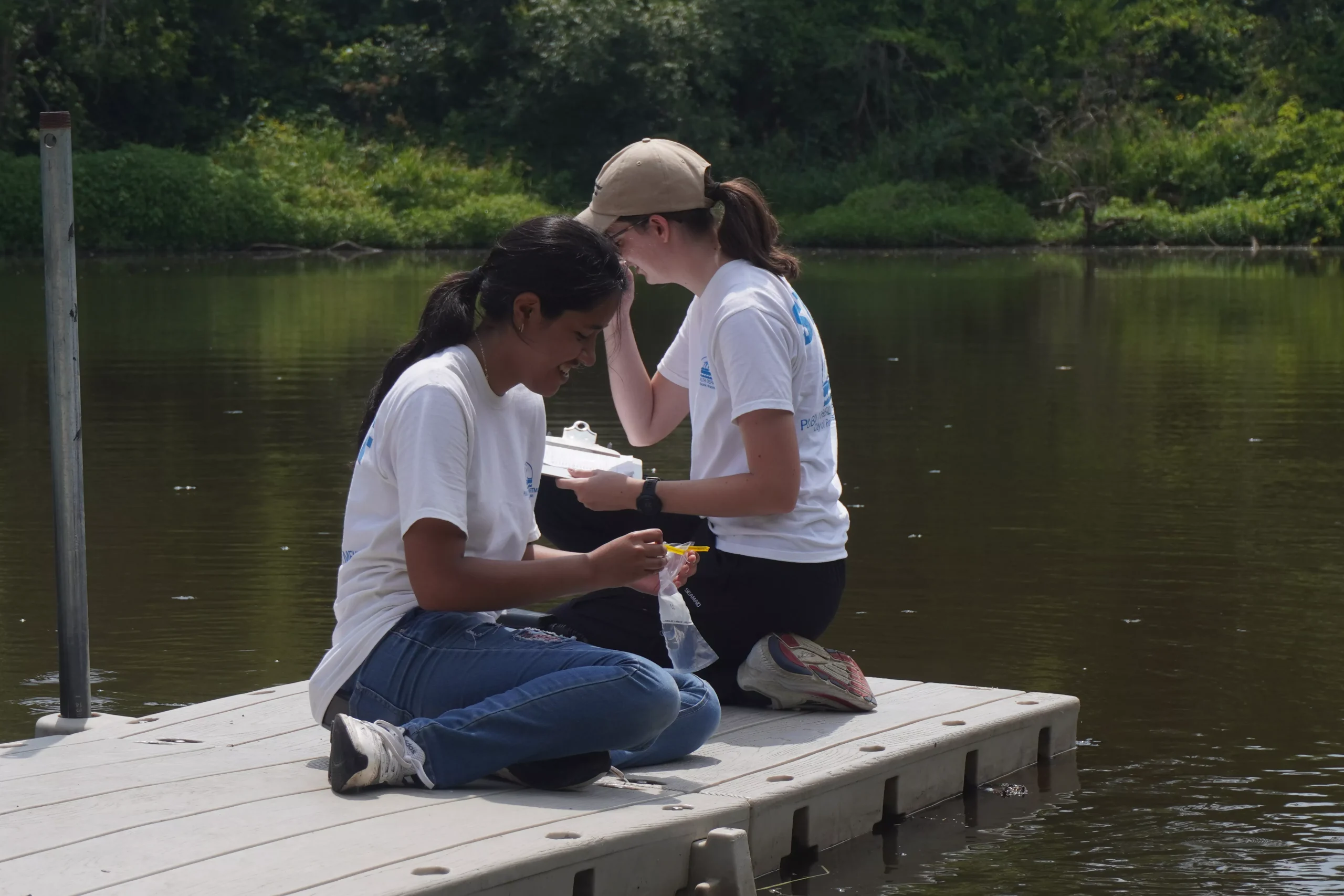 Two women in matching shirts kneeling on a dock, collecting and recording water samples from a lake.