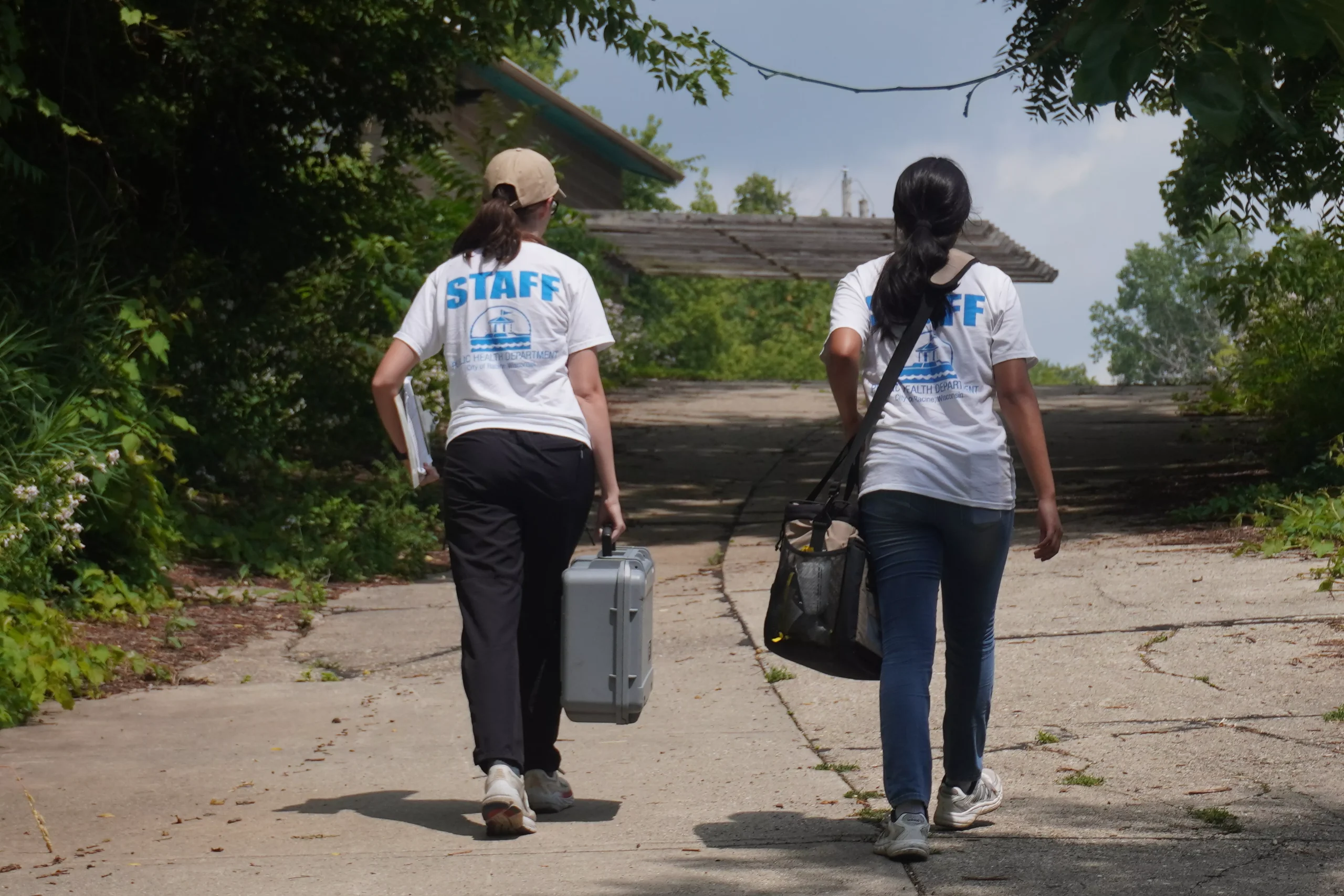 Two staff members walk away on a path, one carrying a case and the other a bag.