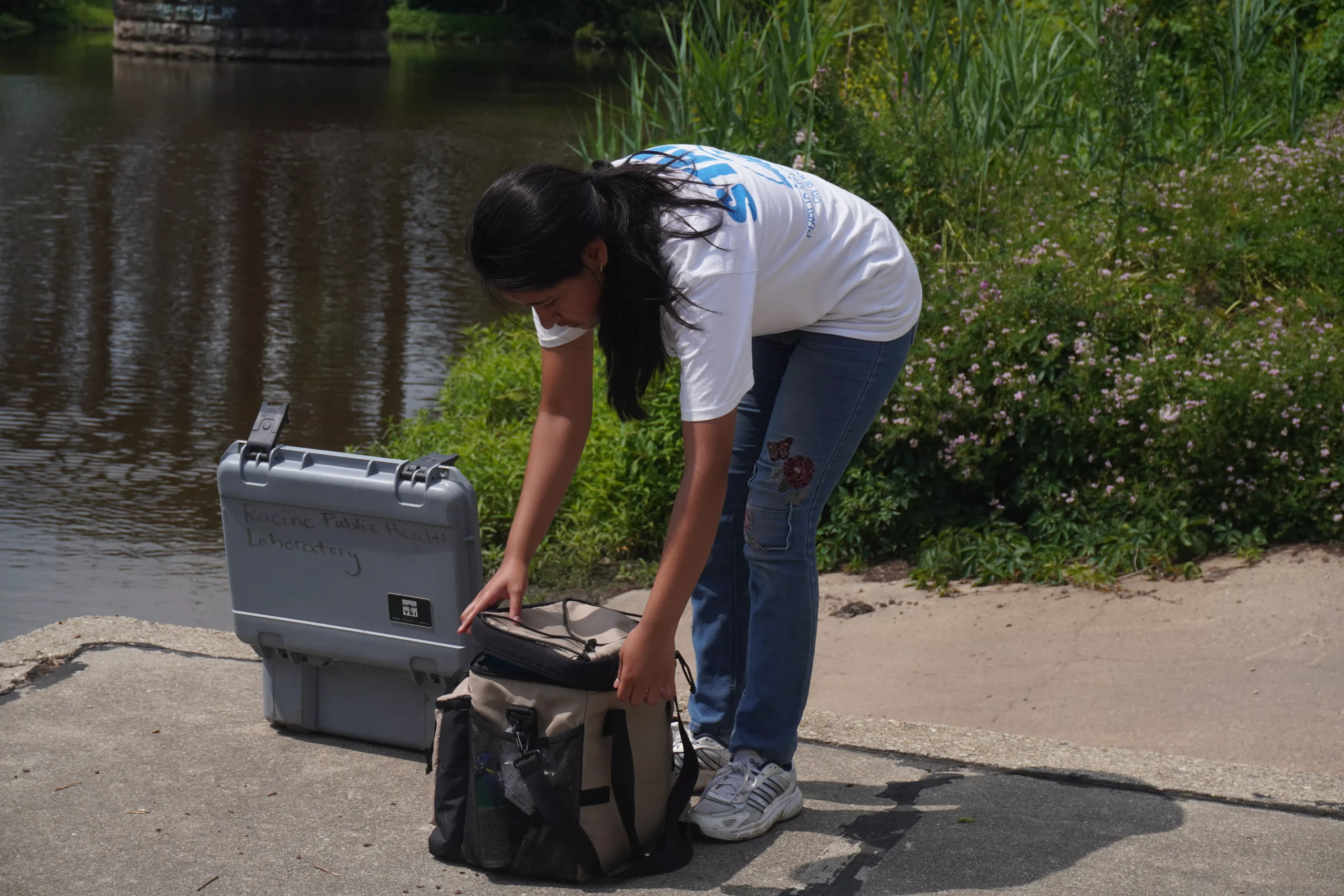 Scientist prepares equipment for water sampling near a river, with a gray case nearby.