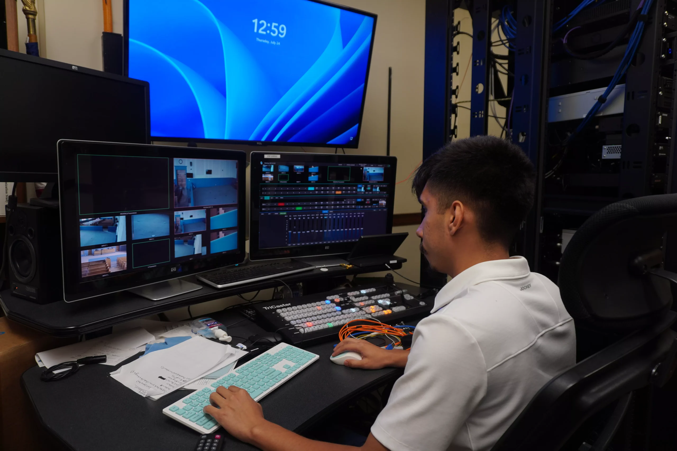 Student edits video in broadcast studio. Multiple monitors display video feeds and editing software interface.
