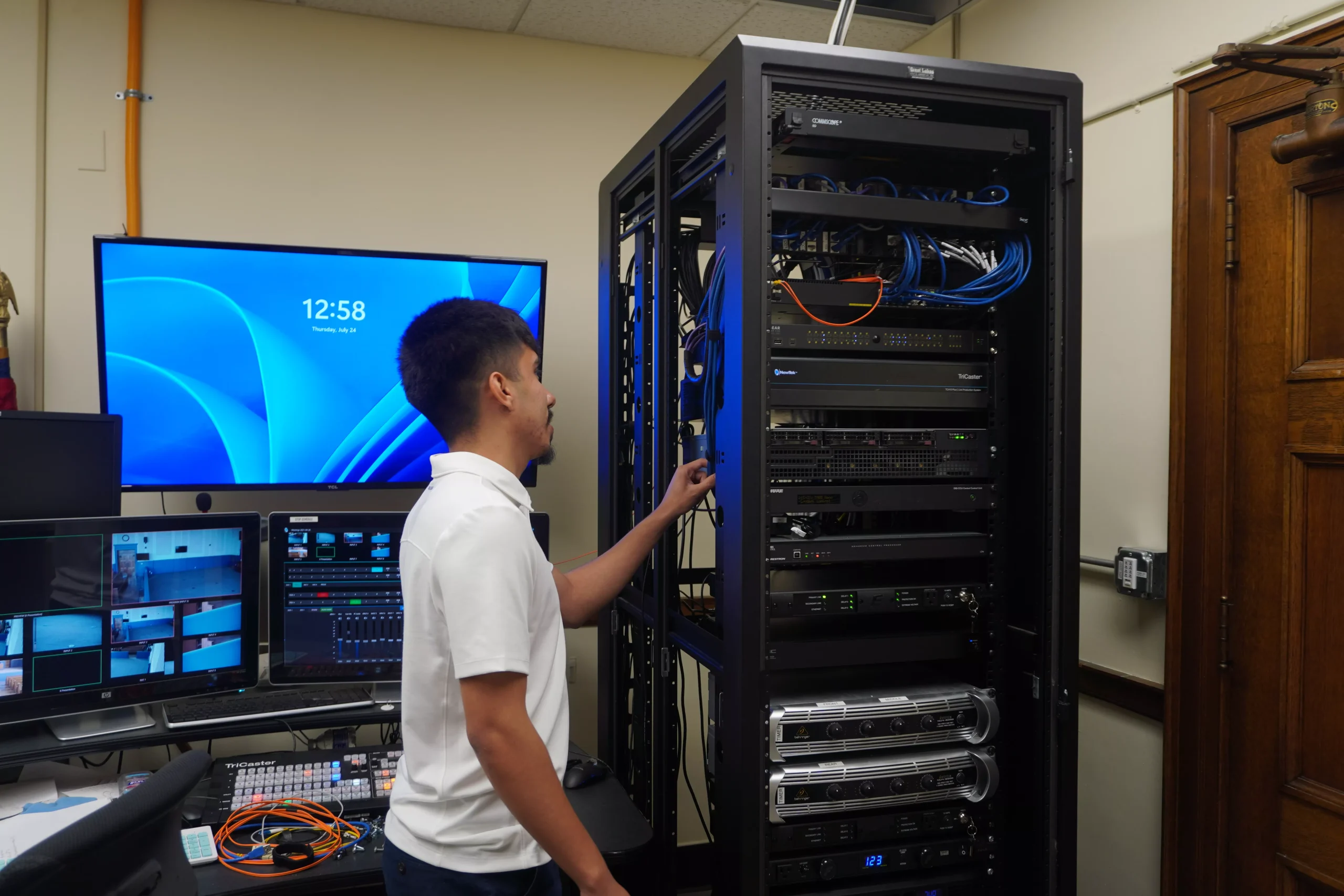 Technician inspecting a server rack in a control room with multiple monitors displaying video feeds.