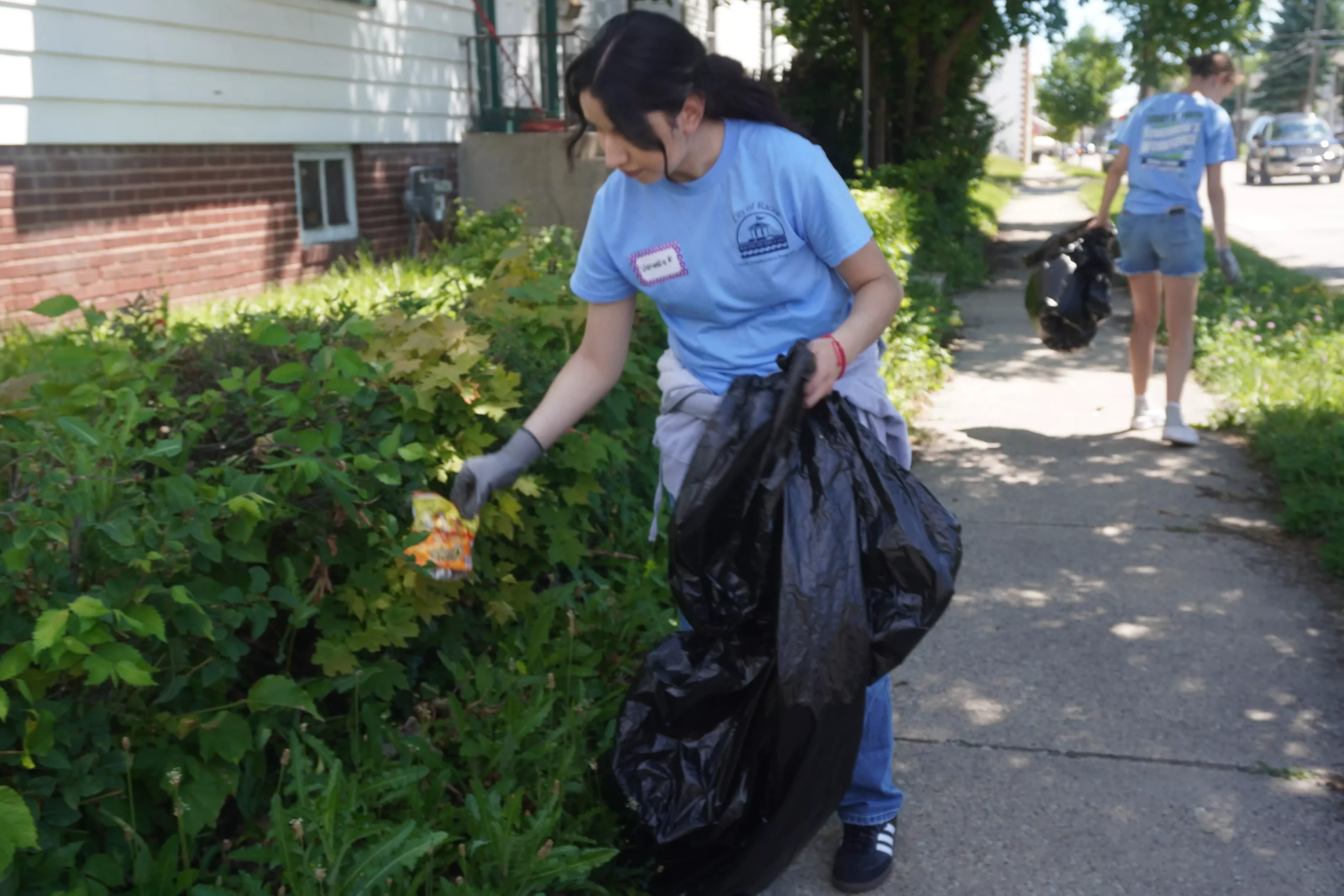 Two volunteers picking up trash along a sidewalk and near bushes, wearing blue shirts and gloves.