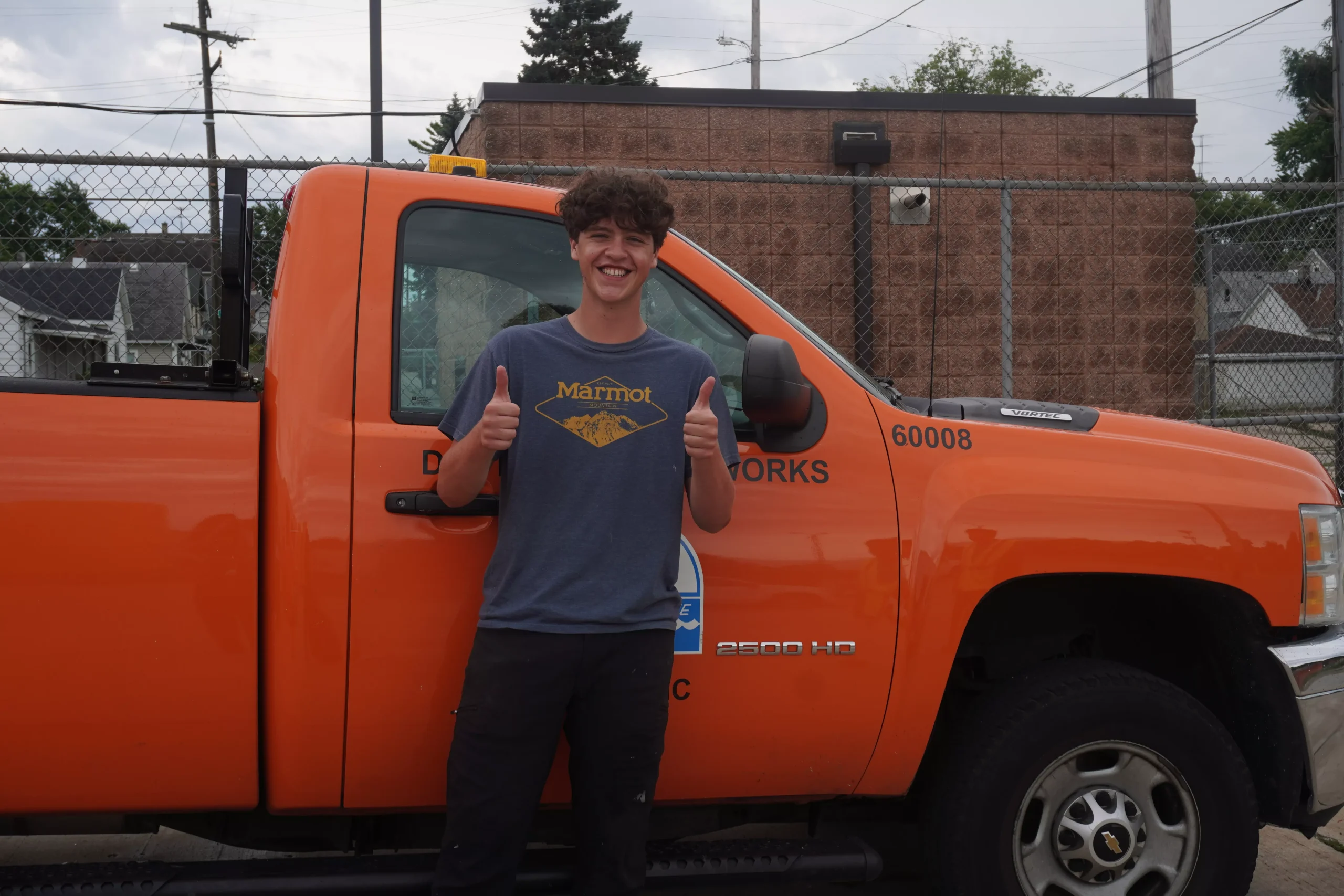 Young man gives thumbs up in front of orange utility truck. Truck has company logo and 'Public Works' on the door.