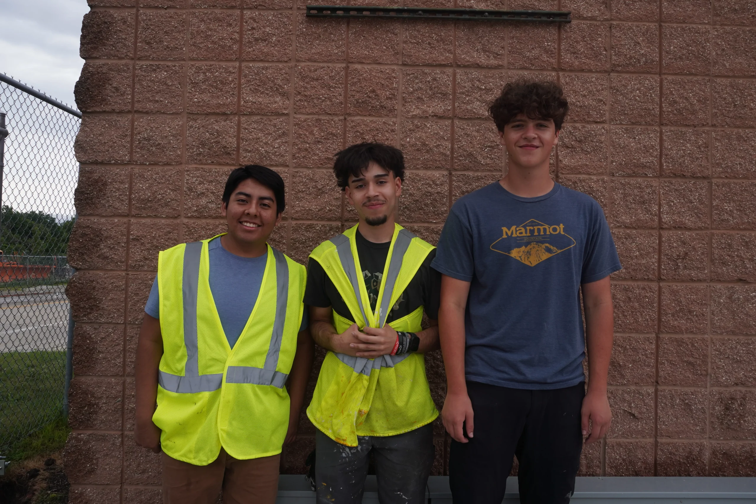 Three young men stand in front of a brick wall, two wearing yellow safety vests.