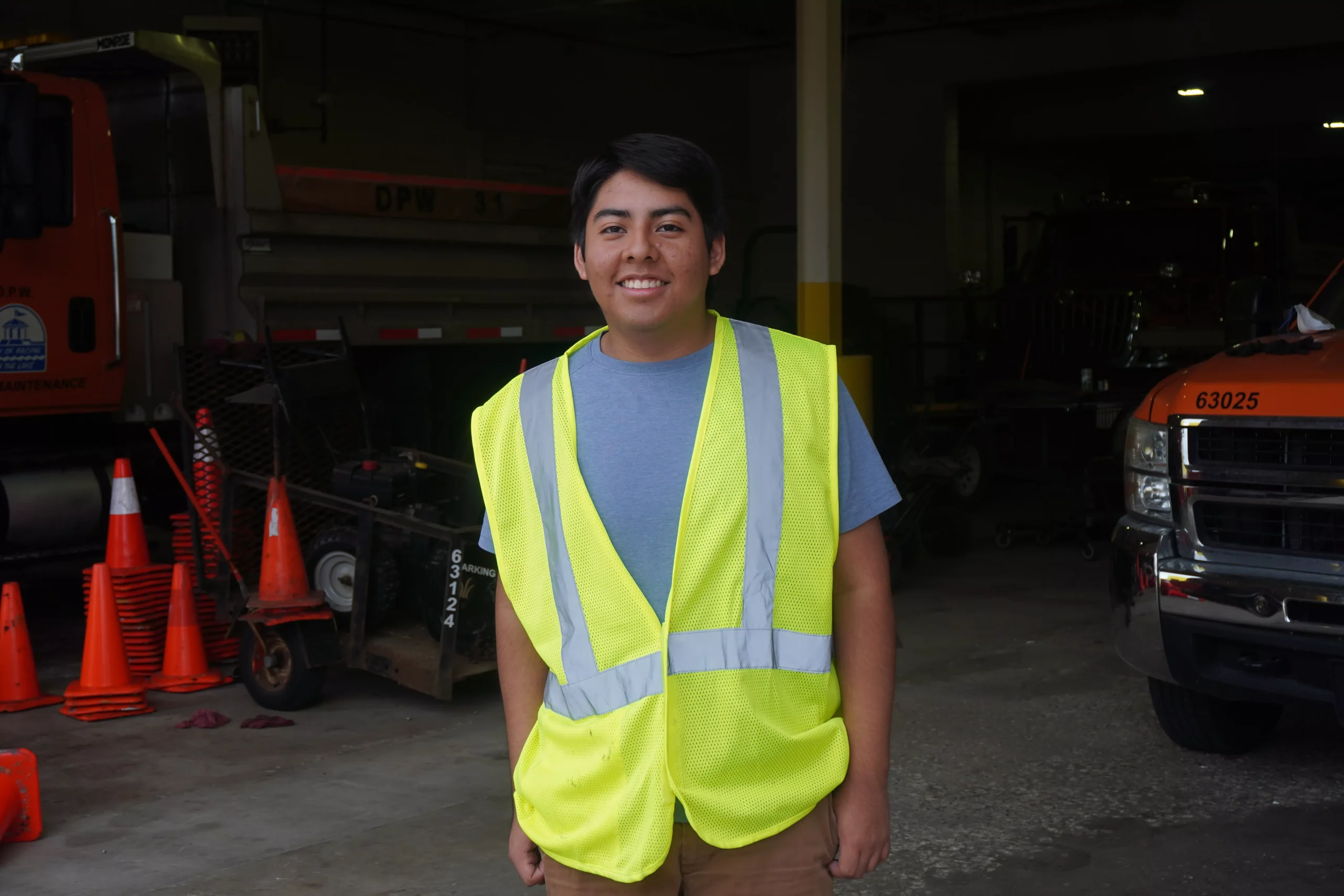 Smiling worker in a safety vest stands in front of a truck and construction equipment.