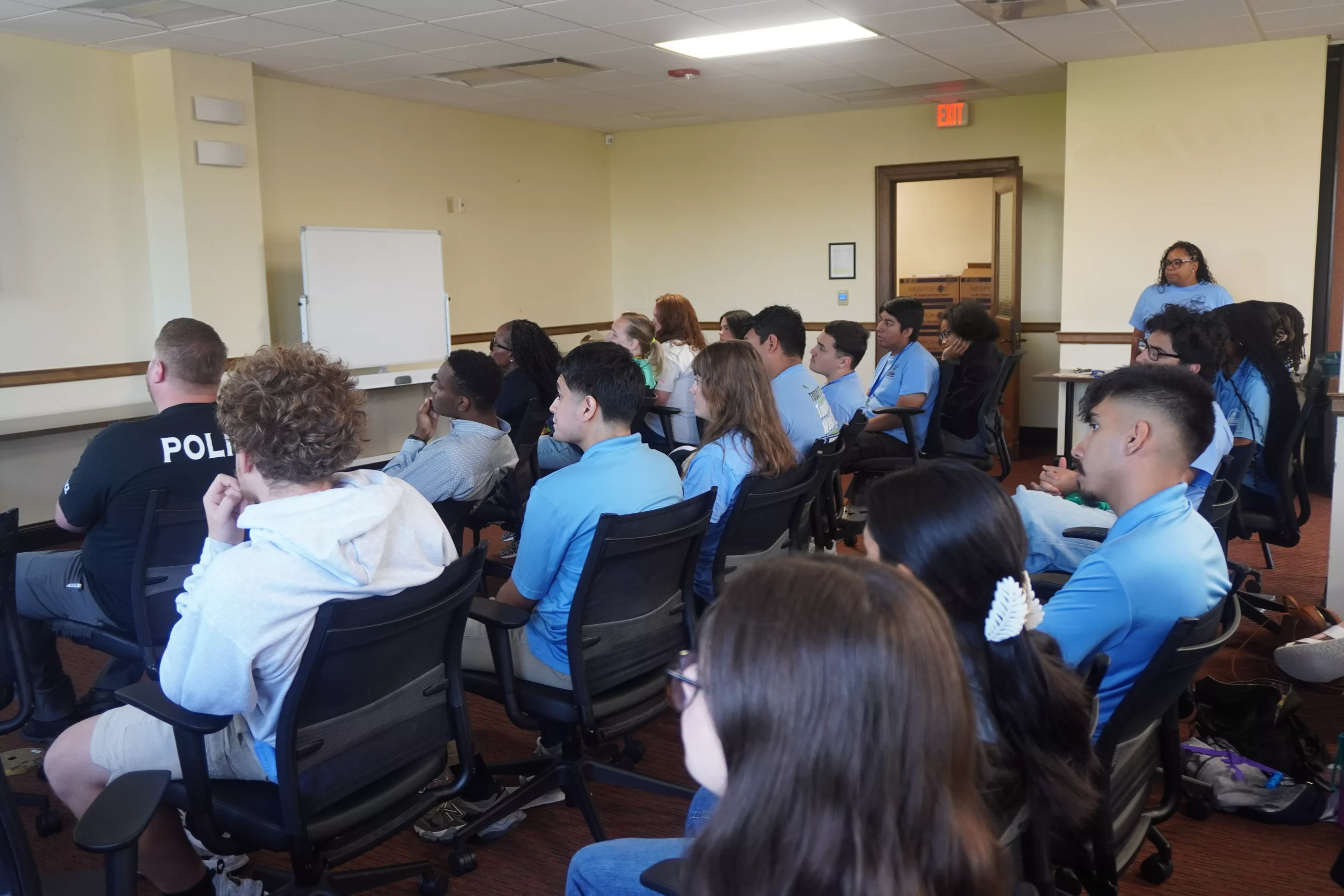 Group of young people seated in a classroom, listening to a presentation. A police officer is also present.